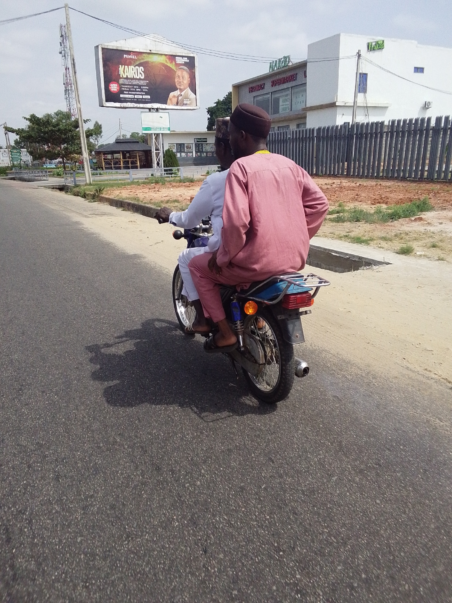Two men are riding on a blue motorcycle down a paved road in Adankolo, Nigeria. The person in the front is wearing white, while the passenger is wearing a pink long-sleeved shirt and pants. They are traveling in the direction of a billboard advertising "KAIROS" by Peniel. In the background is a building with "NAKOD PHARMACY SUPERMARKET" and "Vrakad" written on it. A dark metal fence runs along the edge of a sandy area to the right of the road. A three-legged metal tower is visible behind the billboard. The day appears to be overcast.