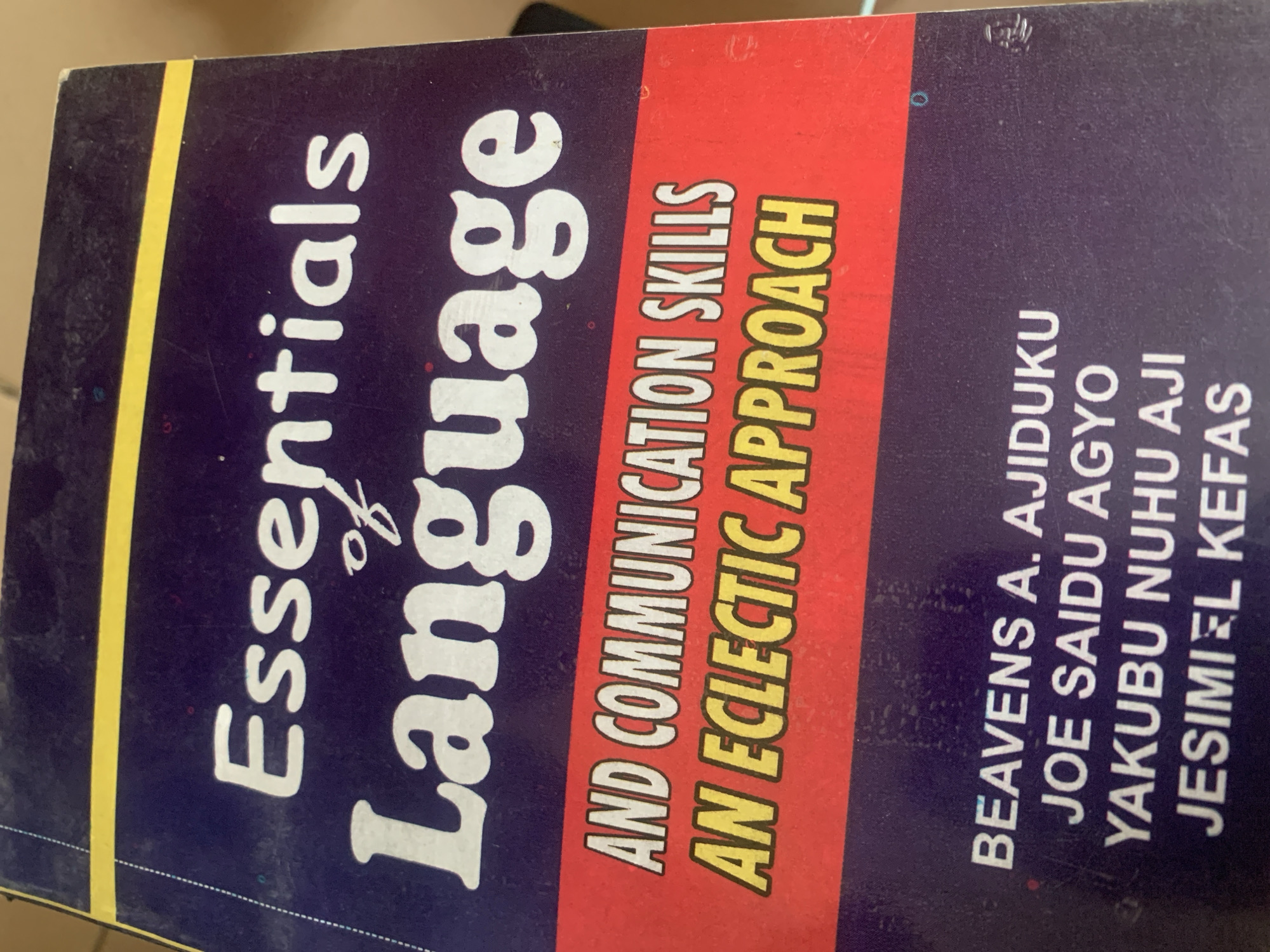 This is a close-up, top-down, eye-level photograph of the cover of a book titled "Essentials of Language and Communication Skills: An Eclectic Approach". The book cover is predominantly dark blue with white and red text. The title "Essentials of Language" is displayed prominently at the top in large white lettering, with "of" in smaller script between "Essentials" and "Language". Below this, the subtitle "AND COMMUNICATION SKILLS" is in white capital letters on a red background, followed by "AN ECLECTIC APPROACH" in yellow capital letters with a black outline, also on the red background. At the bottom of the cover, author names are listed in white capital letters: "BEAVENS A. AJIDUKU", "JOE SAIDU AGYO", "YAKUBU NUHU AJI", and "JESIMIEL KEFAS". The lighting suggests an indoor environment. There are no people, animals, or any indication of the city of Jauro Yinu, Nigeria. The image primarily captures the book's cover, showcasing its title, subtitle, and author information.
