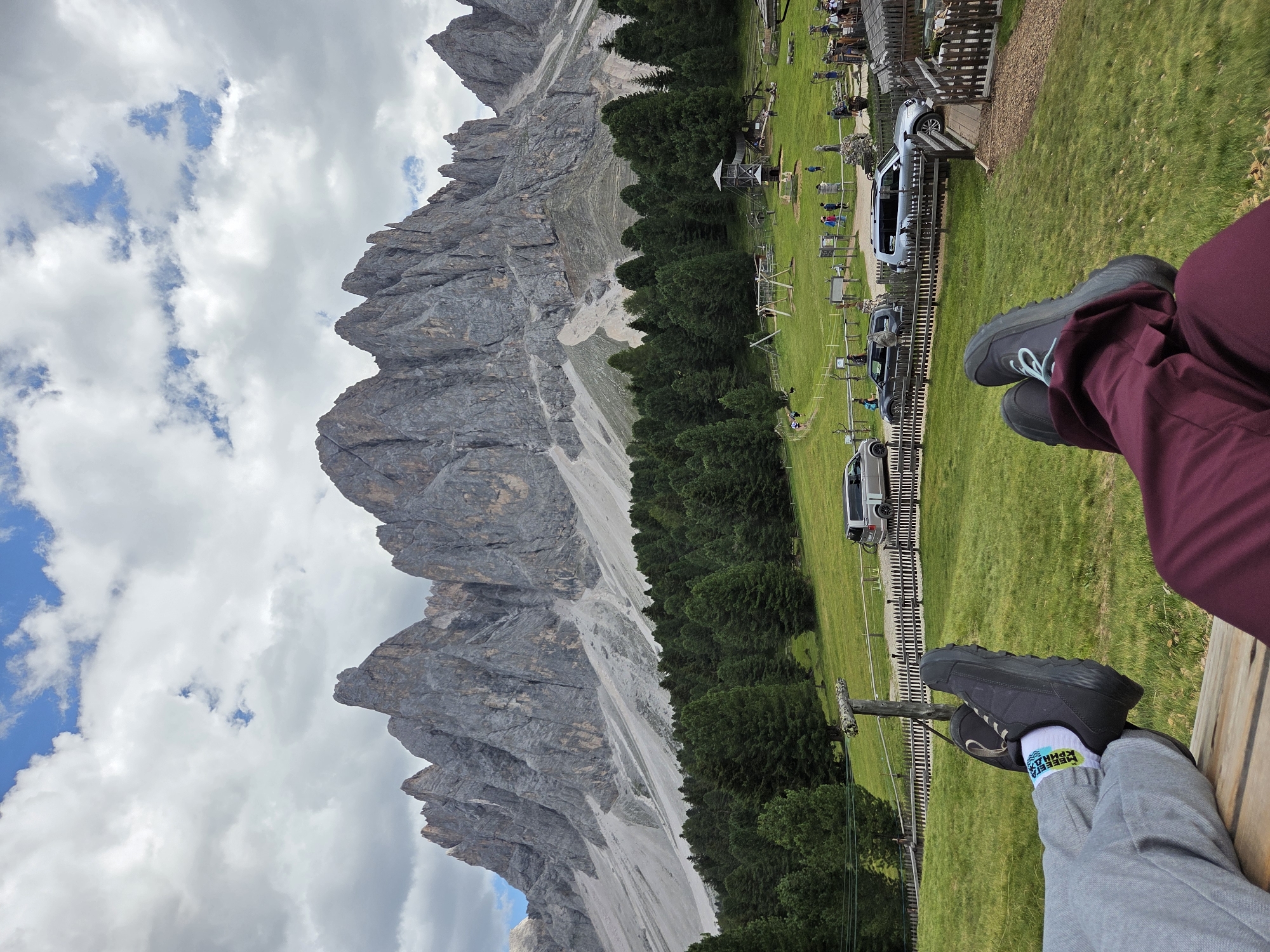 The image shows a scenic view of the Dolomites in Italy. In the foreground, two individuals are seated on a bench, with their legs extended and facing the mountains. The person on the left is wearing grey sweatpants and black athletic shoes with white socks. The person on the right is wearing maroon pants.
The background features a vast expanse of green grass, dotted with trees. Beyond the trees, the majestic Dolomites mountain range rises sharply, with its rugged, rocky peaks piercing a cloudy sky. Several cars are parked in a designated area, and a playground with swings can be seen in the middle ground, indicating this is a popular tourist spot. The overall scene conveys a sense of relaxation and awe in front of the natural beauty of the Italian Alps.