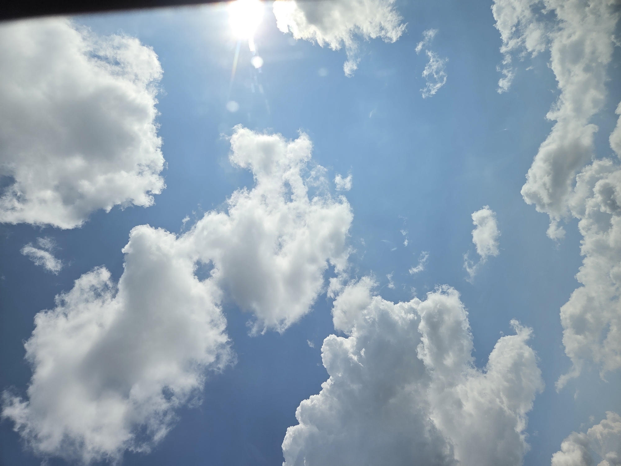 The image is a low-angle shot of a sky in Dalbok Dol, Bulgaria. The sky is blue with scattered cumulus clouds, and the sun is visible as a bright spot at the top of the frame. The clouds are white and fluffy, and they are distributed throughout the frame, occupying the majority of the view. A black object is present in the upper left corner of the frame.