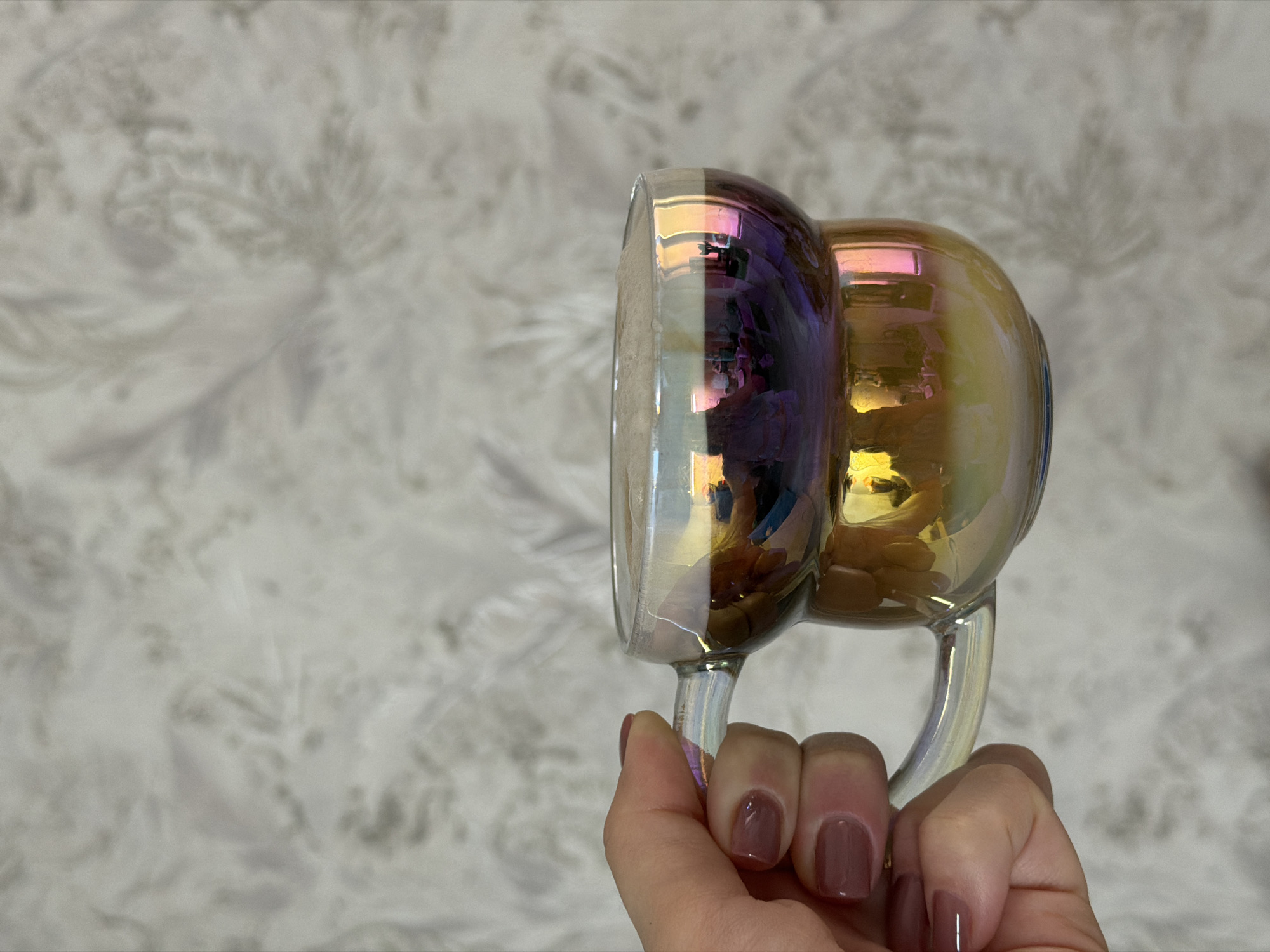 A hand holds a clear glass mug with an iridescent finish, showing purple, gold, and silver hues. The hand has manicured nails with a solid, dark mauve color. The background is a blurred, light gray and white patterned wallpaper. The location appears to be indoors, based on the wallpaper. The image does not provide enough information to pinpoint the location in Reshetnikovo, Russia.