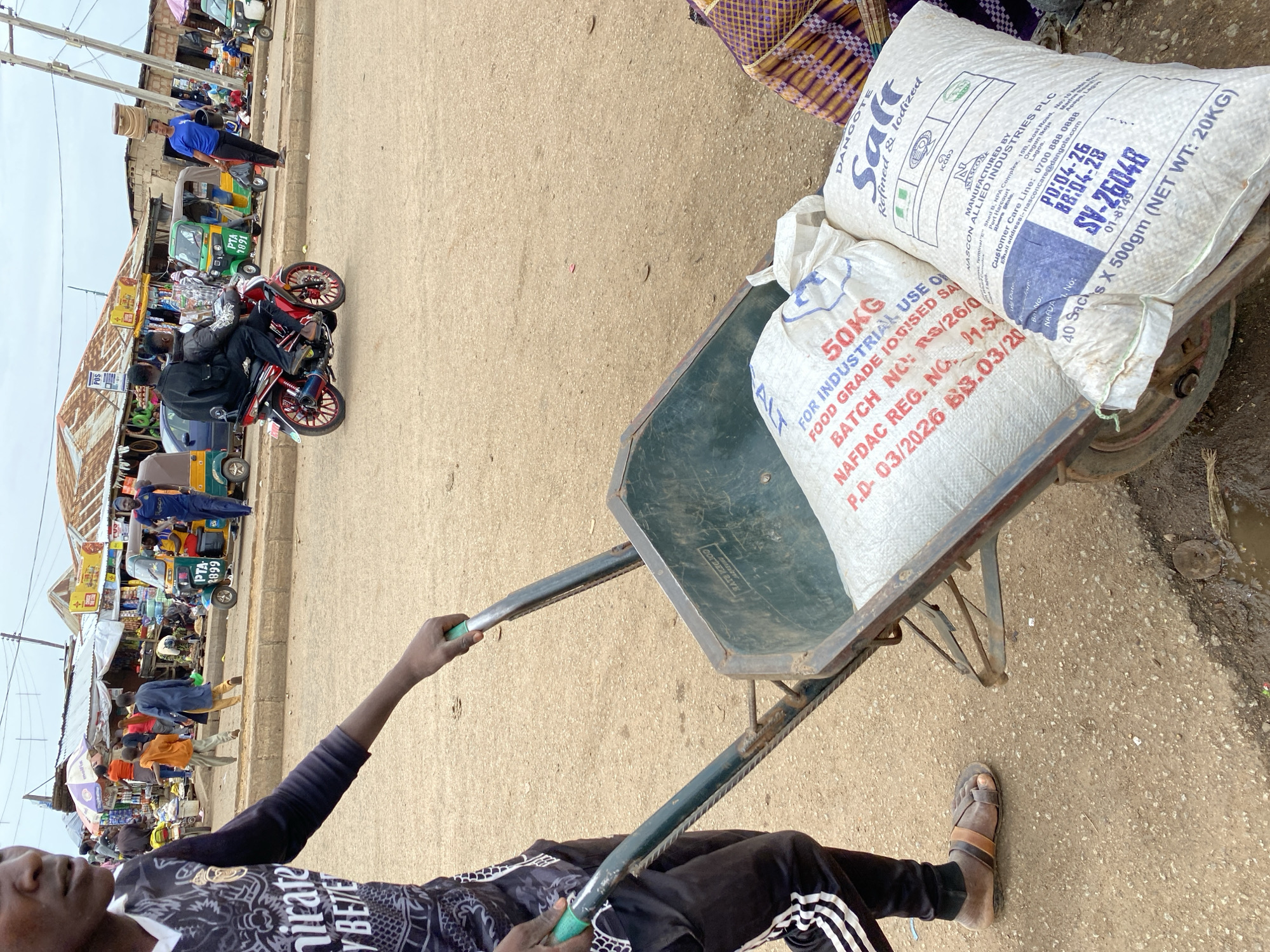 A young male, wearing a black and white jersey, black tracksuit pants with white stripes, and brown sandals, is pushing a green wheelbarrow loaded with two large white sacks on a dirt road in Bukuru, Nigeria. The sack on the left has red text that reads "50KG FOR INDUSTRIAL USE ONLY FOOD GRADE IODISED SAL," along with batch and registration numbers. The sack on the right, labeled "DANGOTE Salt Refined & Iodized," contains 20kg of salt. In the background, there is a bustling marketplace with various stalls, people walking, and three-wheeled auto-rickshaws (keke napep). A red motorcycle with two passengers is also visible in the middle ground. The sky is overcast.