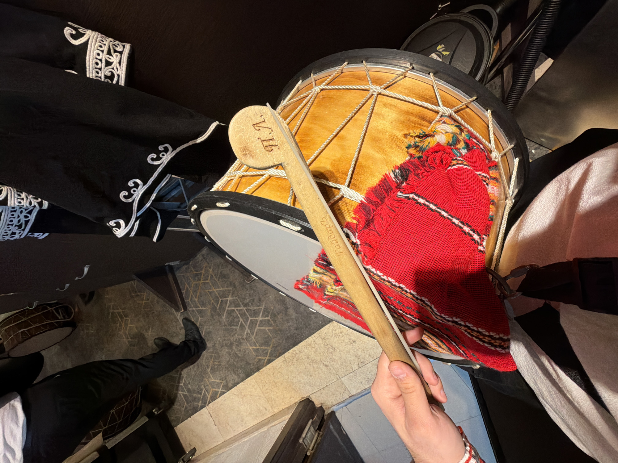 A hand holds a wooden drumstick (labeled "gadułka") with the letters "VI" branded on the head, positioned above a tupan drum draped with a red patterned textile. The drum, which is wooden with rope tensioners, sits among various items including what appears to be a black garment with embroidered white trim, a white garment, and other black objects. A person wearing black pants and black shoes is partially visible in the lower left of the frame, suggesting they are standing near the drum. The image appears to be taken indoors, potentially at ground level. The presence of traditional Bulgarian musical instruments suggests the location is in Varna, Bulgaria.