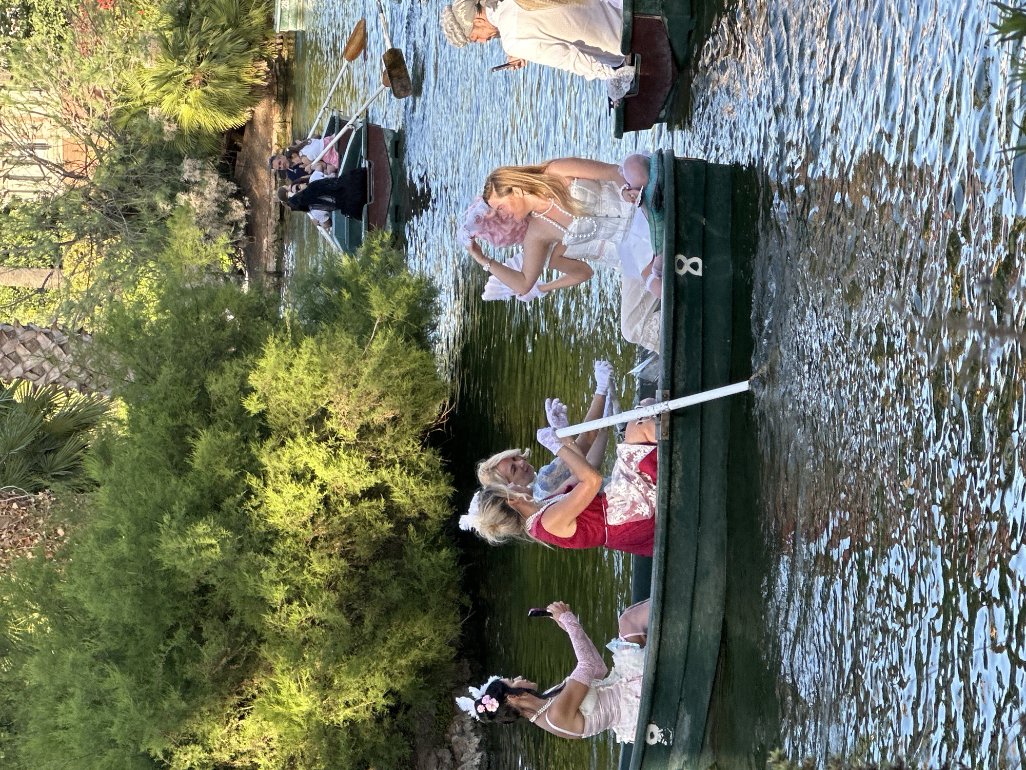 This detailed image captures a vibrant scene of individuals in costumed attire enjoying rowing boats on a lake in Parc de la Ciutadella, Barcelona, Spain. The setting is an open-air park environment characterized by lush, varied green foliage, including dense bushes and taller trees, with distinct palm trees visible among them. In the far background, behind the trees, a light-colored building with a dark roofline can be discerned, consistent with structures found within the park. The water in the lake is a deep green hue, reflecting the surrounding greenery, with ripples on its surface indicating gentle movement. The lighting suggests a warm, bright day, possibly late afternoon, with sunlight casting highlights on the water and foliage. In the foreground, two dark green wooden rowing boats are prominent, alongside other boats in the mid-ground. The leftmost boat, numbered "8" in white on its bow, contains three women. The woman at the stern, with dark hair, faces right and holds a dark-colored smartphone vertically, appearing to capture a photo or video. She wears a light pink, layered outfit with a corset-style top, a pearl necklace, and long, light pink fingerless gloves. Her hair is adorned with white and pink accessories. The woman in the middle, with blonde hair, actively rows an oar with a white blade, facing forward. She wears a red dress with a white lace apron overlay, a pearl necklace, and long, light purple or white gloves. The third woman, at the bow, also blonde, faces forward and slightly left, looking towards the middle rower. She wears a light blue or white dress and light purple or white gloves. Adjacent to the right, another partially visible dark green/brown rowing boat holds two individuals. A blonde woman at the stern faces forward and slightly left, holding a pink, feathery object (possibly a boa or fan) near her face. She is dressed in a white corset-style top, a pearl necklace, white flowing trousers or a skirt, and light pink slippers or flat shoes. Beside her, a man at the bow, sporting a white, period-style wig, faces forward and slightly left, also holding a dark-colored smartphone vertically, seemingly taking a photo or video. He wears a white button-up shirt. In the mid-ground, a dark-colored boat with two individuals rowing and two seated passengers is visible. Further back and to the right, another partially visible dark boat also contains occupants and oars. The scene