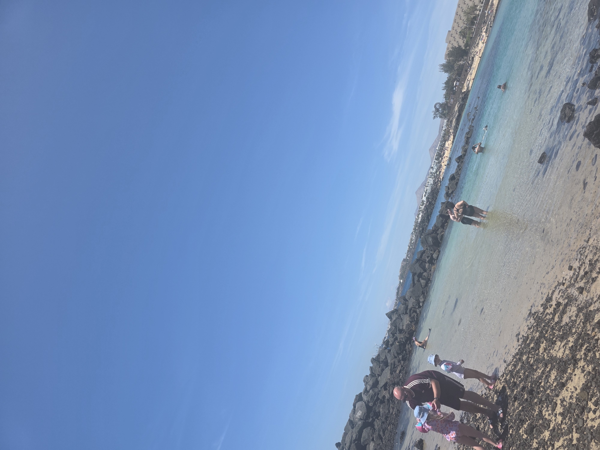 A medium shot captures a sunny day at a beach in Costa Teguise, Spain. The foreground is dominated by dark volcanic rocks and a sandy shore with shallow, clear blue water. Several people are visible. On the left, a man in a burgundy t-shirt and dark shorts stands with two young girls, one in a pink swimsuit and blue hat, the other in a white swimsuit and blue hat. The man appears to be holding the hand of the girl on the left. In the water, a couple stands partially submerged. Further out, a person is lying on a bodyboard, and two other individuals are swimming. The middle ground shows a calm, turquoise bay with more swimmers. The background reveals a resort town with buildings and palm trees lining the coastline, with hills in the distance under a bright blue sky with sparse clouds.