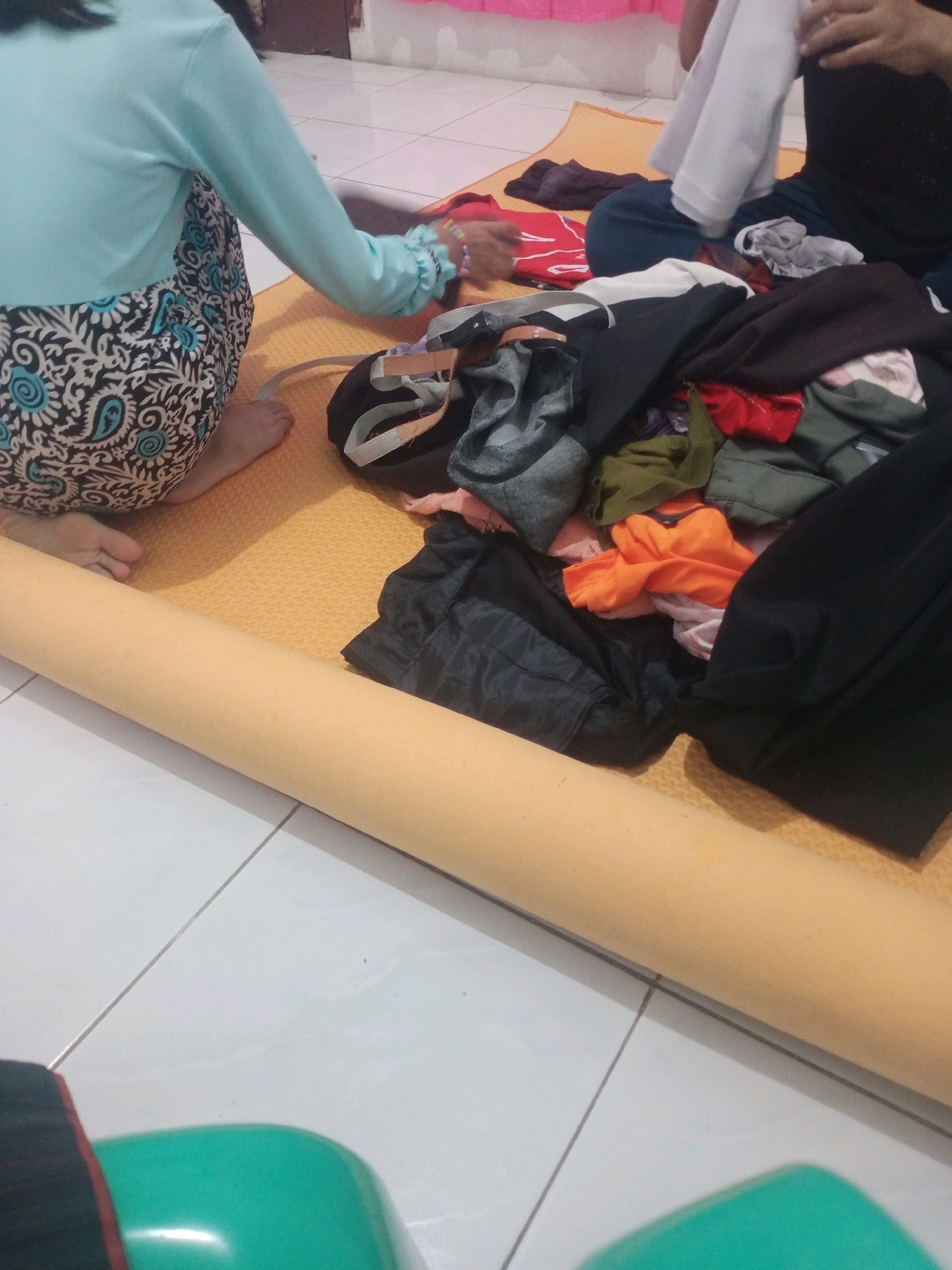 In a room with tiled flooring, a light beige yoga mat is laid out. On the mat, a pile of assorted clothing is arranged, including dark-colored garments, an orange shirt, and a white shirt. Two individuals are partially visible. One, wearing a light blue long-sleeved shirt and a patterned skirt, is seated on the mat. Another person, dressed in black, is also seated, holding a white piece of fabric. A large, cylindrical object, possibly a rolled-up mat or carpet, lies horizontally across the foreground. The environment appears to be domestic, possibly a living room or bedroom, within the city of Bandar Lampung, Indonesia.