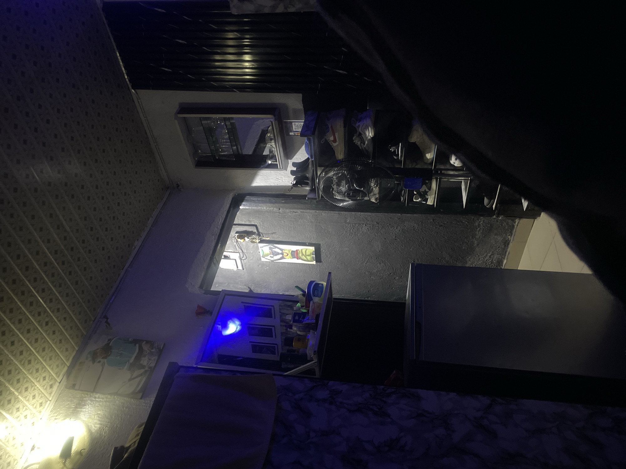 The image depicts an indoor scene, possibly a bedroom or living area, with low lighting and a cool blue hue emanating from a light source in the upper left corner. A poster of a person in a blue sweatshirt is mounted on the wall above a shelf containing various items, including bottles and containers. To the right, a shoe rack holds several pairs of sneakers. A tall, silver appliance, likely a refrigerator, is positioned to the left. The walls appear to be painted a dark gray, and the ceiling has a textured, tiled pattern. The overall impression is that of a cluttered but lived-in space.