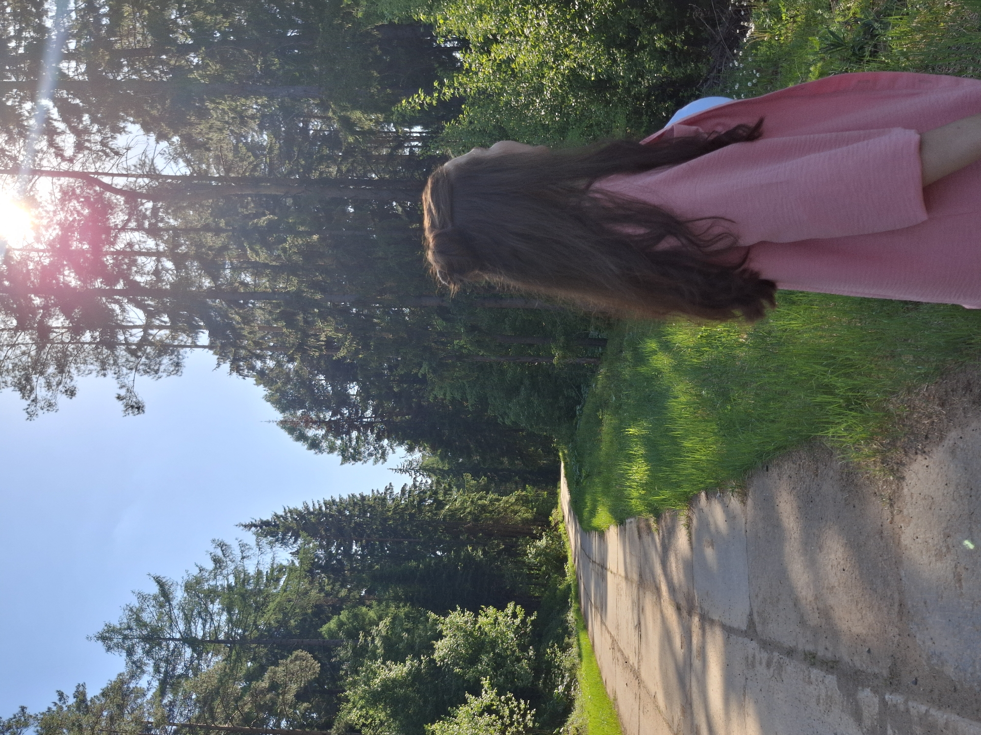 The image depicts a woman with long brown hair, wearing a pink dress, standing on a concrete path in a wooded area. The path runs through tall green trees under a clear blue sky with the sun shining brightly. The location appears to be in Прикамский, Russia.