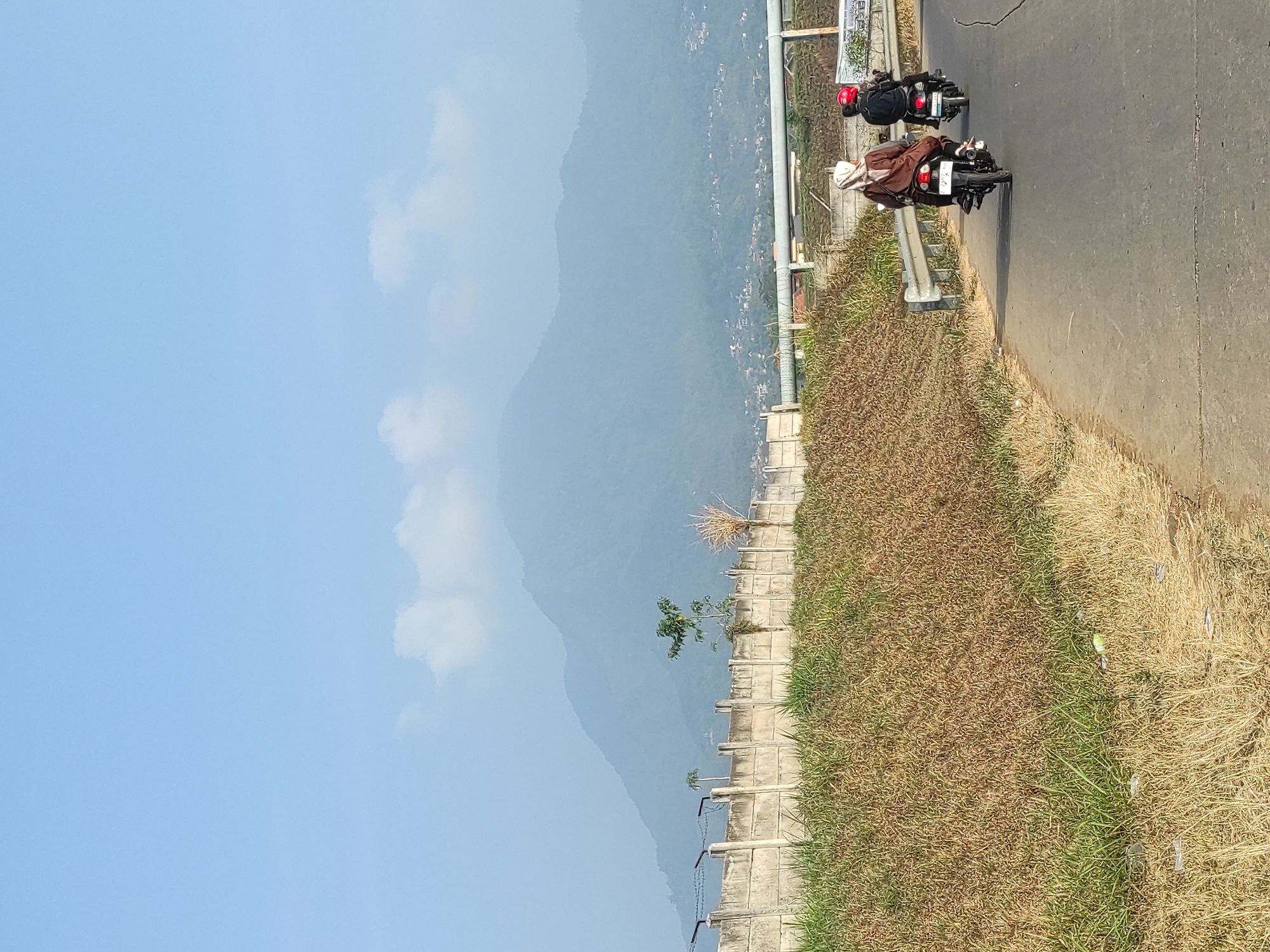 In Sumedang, Indonesia, a two-lane asphalt road curves slightly to the right. Two individuals on motorcycles are traveling away from the viewer. The person in front is wearing a brown headscarf and a brown garment, riding a black motorcycle. Behind them, a person wearing a red helmet and a black jacket is on another black motorcycle. The road is bordered on the left by a dry, grassy embankment that leads up to a concrete wall with barbed wire on top. Beyond the wall, a sparse tree grows. In the background, a hazy, green mountain range dominates the landscape under a clear blue sky with a few wispy clouds. Some structures are visible on the slopes of the mountains. A metal guardrail runs along the right side of the road.