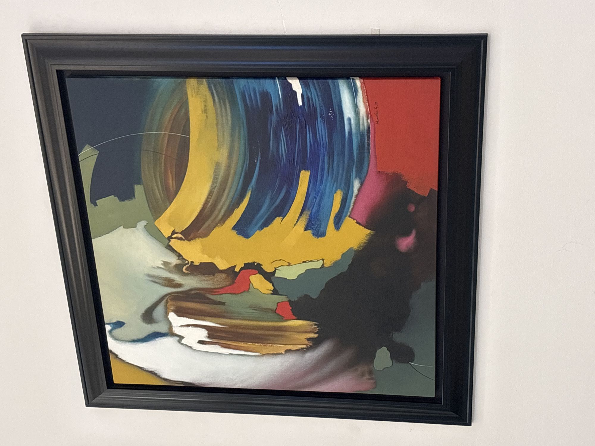 A framed abstract painting hangs on a white wall. The painting features bold brushstrokes and a vibrant color palette, dominated by yellows, blues, and reds, with areas of white, brown, and black. The frame is thick and black. The painting appears to be signed in the lower right corner with "Marden G. 19". The location of this image is not provided in the prompt.