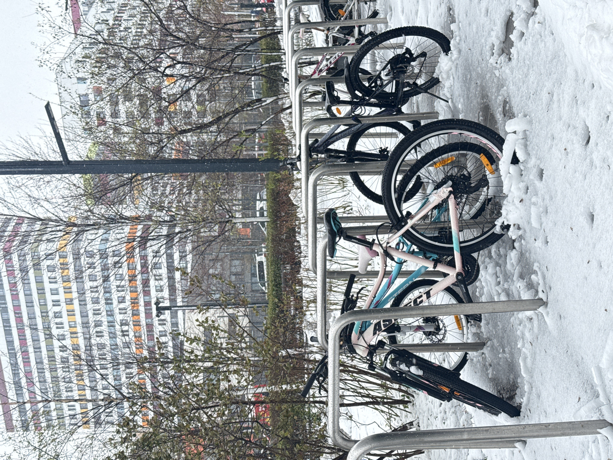 The image captures an urban winter scene in Moscow, Russia, after a fresh snowfall. In the foreground, several bicycles are parked upright in a sturdy, metallic bike rack. The ground around the rack and parts of the bicycles are covered in a layer of soft, white snow, suggesting cold weather. Snow also clings to the bare branches of deciduous trees and some green bushes that are scattered throughout the mid-ground.
One bicycle prominently features in the center, identifiable by its light blue and pink frame and white tires. Visible text on its frame includes "STERN" and "CT," likely part of a brand name like "CONNECT." This bike is equipped with disc brakes and orange reflectors on its wheels. Other bicycles in the rack are less distinct, appearing mostly dark-colored.
In the background, towering multi-story buildings dominate the skyline, showcasing numerous windows with some frames or panels featuring distinct colors such as orange, red, blue, and yellow. These structures appear to be modern residential or office blocks, typical of urban architecture. A tall, dark pole, possibly a streetlight or utility pole, stands on the left side of the image.
The scene appears to be set during daytime under an overcast sky, providing diffused light that highlights the fresh snow. There is no human activity visible, creating a quiet and somewhat still atmosphere. The fresh snow suggests a recent snowfall, contributing to the distinct winter aesthetic.
