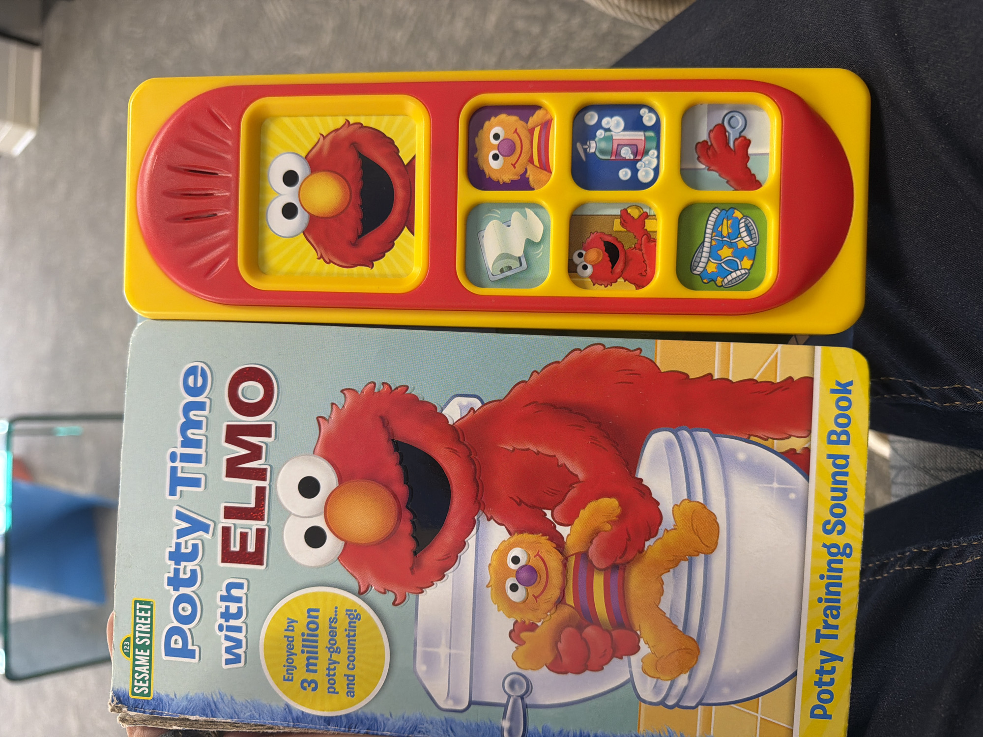 A "Potty Time with Elmo" sound book is displayed next to a yellow and red Elmo-themed electronic toy. The sound book features an illustration of Elmo and a character named "Grover" sitting on a toilet. The electronic toy has a red curved top and displays six small squares with images of Elmo characters and related objects, such as toilet paper and soap. The book's cover also advertises "Sesame Street" and mentions that it is "enjoyed by 3 million potty-goers... and counting!". The scene appears to be indoors, possibly in a home or classroom environment, but the specific location in Chesapeake, United States of America cannot be determined from the image.