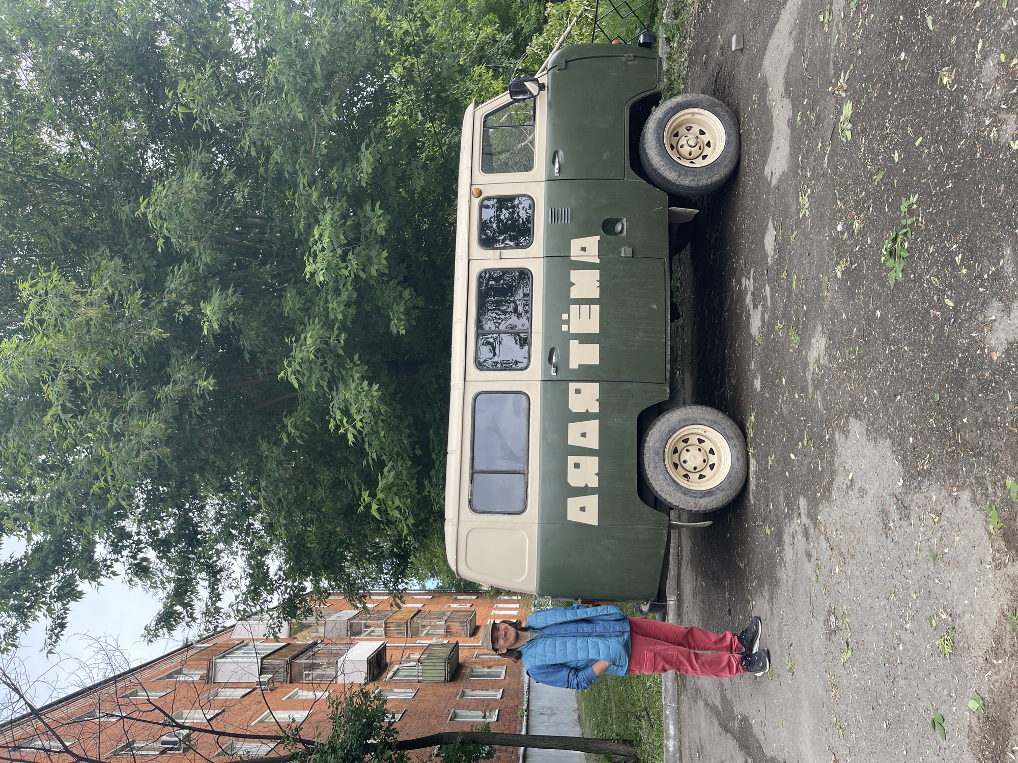 A person in a blue jacket and red pants stands next to a two-toned, beige and green UAZ-452 van with Cyrillic writing on its side that appears to read "Artyom" in Russian. The person faces the camera, smiling slightly. The van is parked on a worn asphalt surface with scattered leaves and patches of dampness. Behind the van and person, there are trees and a brick building. The overall scene suggests a location in Izhevsk, Russia.