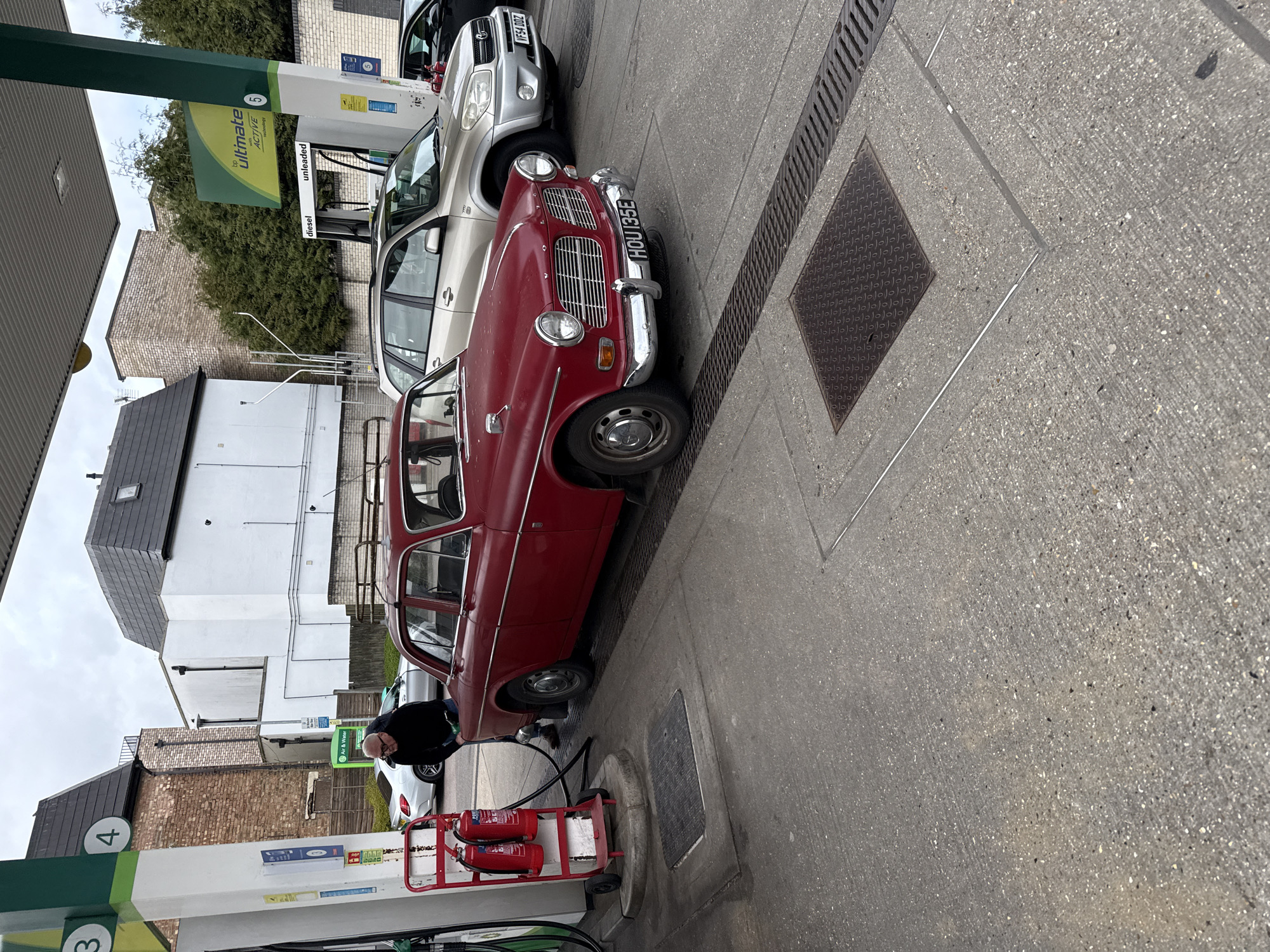 The image captures a scene at a BP petrol station. The red vintage car, with the license plate "HOU 135E", is parked at a pump. A silver SUV, bearing the license plate "VF54 DDZ," is parked adjacent to it. An individual in a black top is refueling the car. The BP fuel pump is labeled "4", and indicates "Air & Water". The sign overhead denotes "Ultimate ACTIVE 5" with unleaded and diesel options. Adjacent to the pump is a red trolley with two fire extinguishers. The surroundings include buildings with brick and white facades, and a black tiled roof.