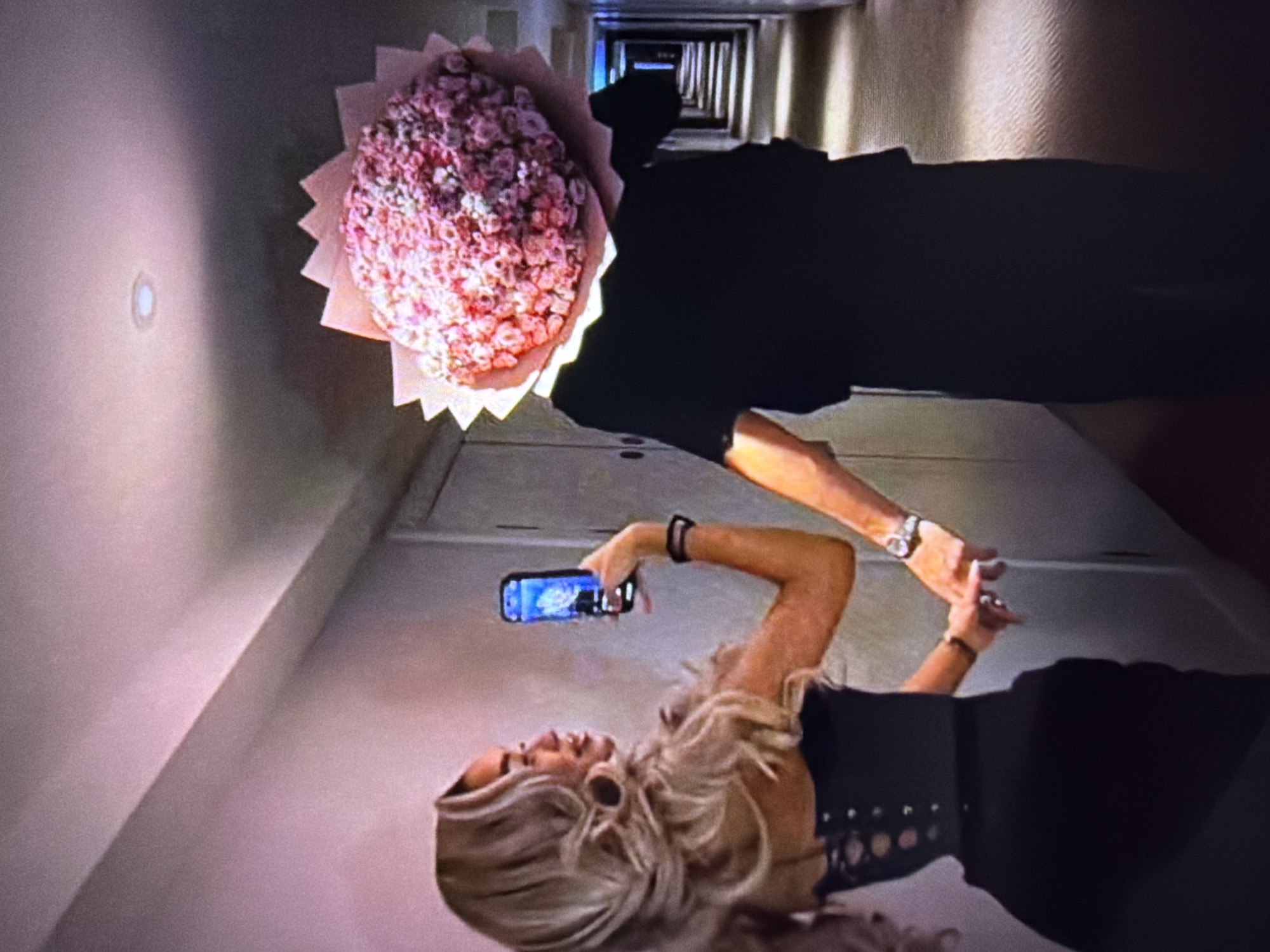 The image depicts two individuals in a dimly lit, narrow hallway. The hallway appears to be made of concrete or similar material, with a textured pattern on the walls. A series of identical doors or openings are visible down the length of the corridor, suggesting a long, uniform structure like a hotel or apartment building hallway.
In the foreground, a woman with blonde, wavy hair is holding a smartphone horizontally, seemingly taking a picture or recording a video. She is wearing a black, sleeveless dress with a corset-like design. A watch is visible on her left wrist. Her posture suggests she is looking at the phone's screen.
Behind her and slightly to the left, a person in a dark outfit, possibly a suit, is holding a large bouquet of pink flowers wrapped in light pink paper with a serrated edge. The bouquet is positioned in front of the person's torso. The flowers are small and numerous, giving the bouquet a dense, rounded appearance. The person's body is partially obscured by the bouquet.
The lighting in the scene is subdued, with a single light fixture visible on the wall to the left, casting a soft glow. The overall atmosphere is somewhat surreal and potentially intimate, given the close proximity of the individuals and the presence of the flowers. The perspective is unusual, as if the camera is angled upwards or the subjects are positioned in a way that creates this effect. The time of day is not discernible, and there is no indication of weather. No text is visible in the image.