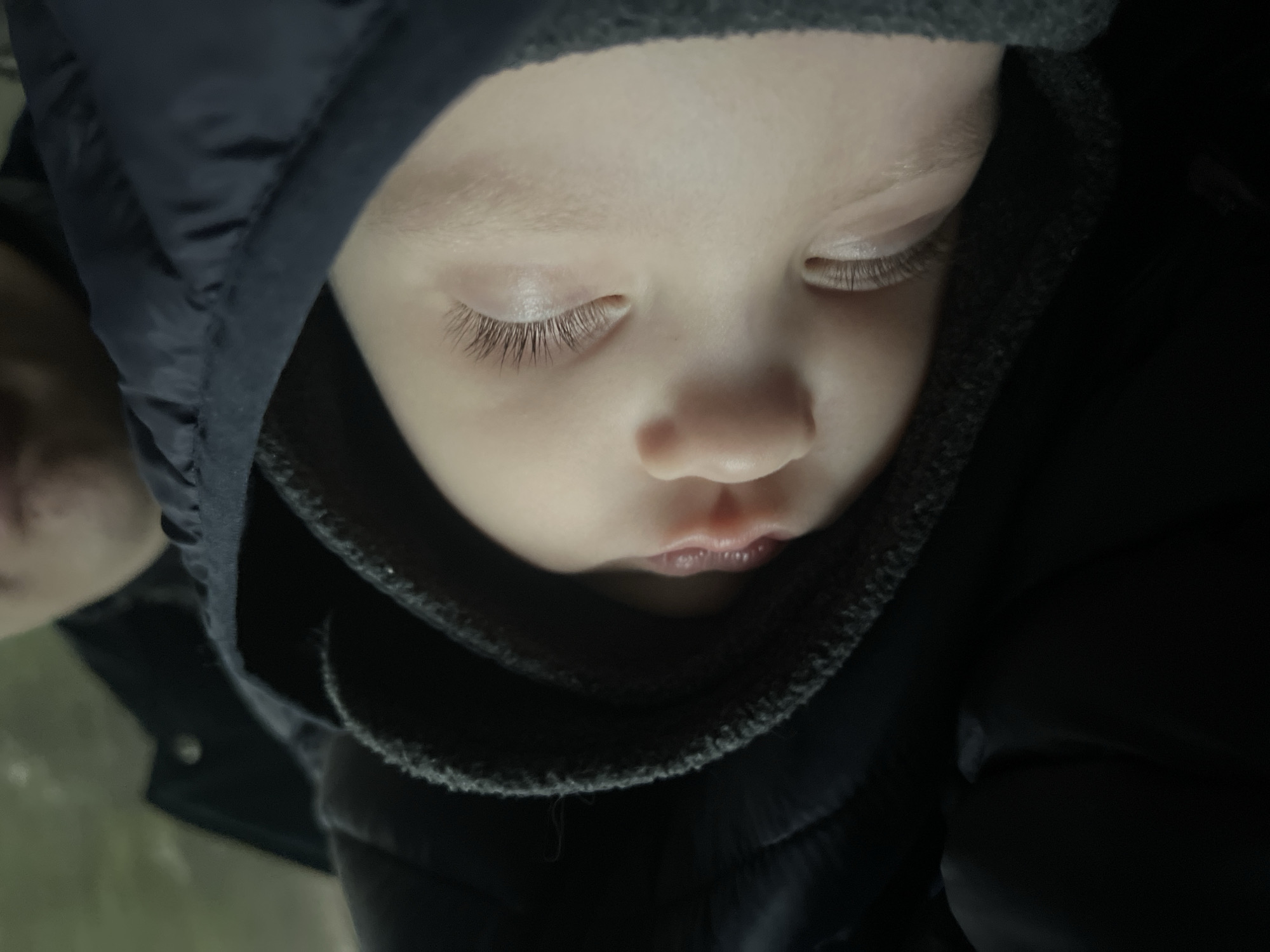 A close-up, downward-angled shot shows a child's face. The child has pale skin, closed eyes, and long eyelashes. The child is wearing a dark-colored, hooded jacket or coat, and a dark gray scarf or garment is around their neck. Part of an adult's face is visible on the left side of the frame. The lighting is soft, and the background is dark and indistinct. The image does not contain any information suggesting it was taken in Chaikovsky, Russia.