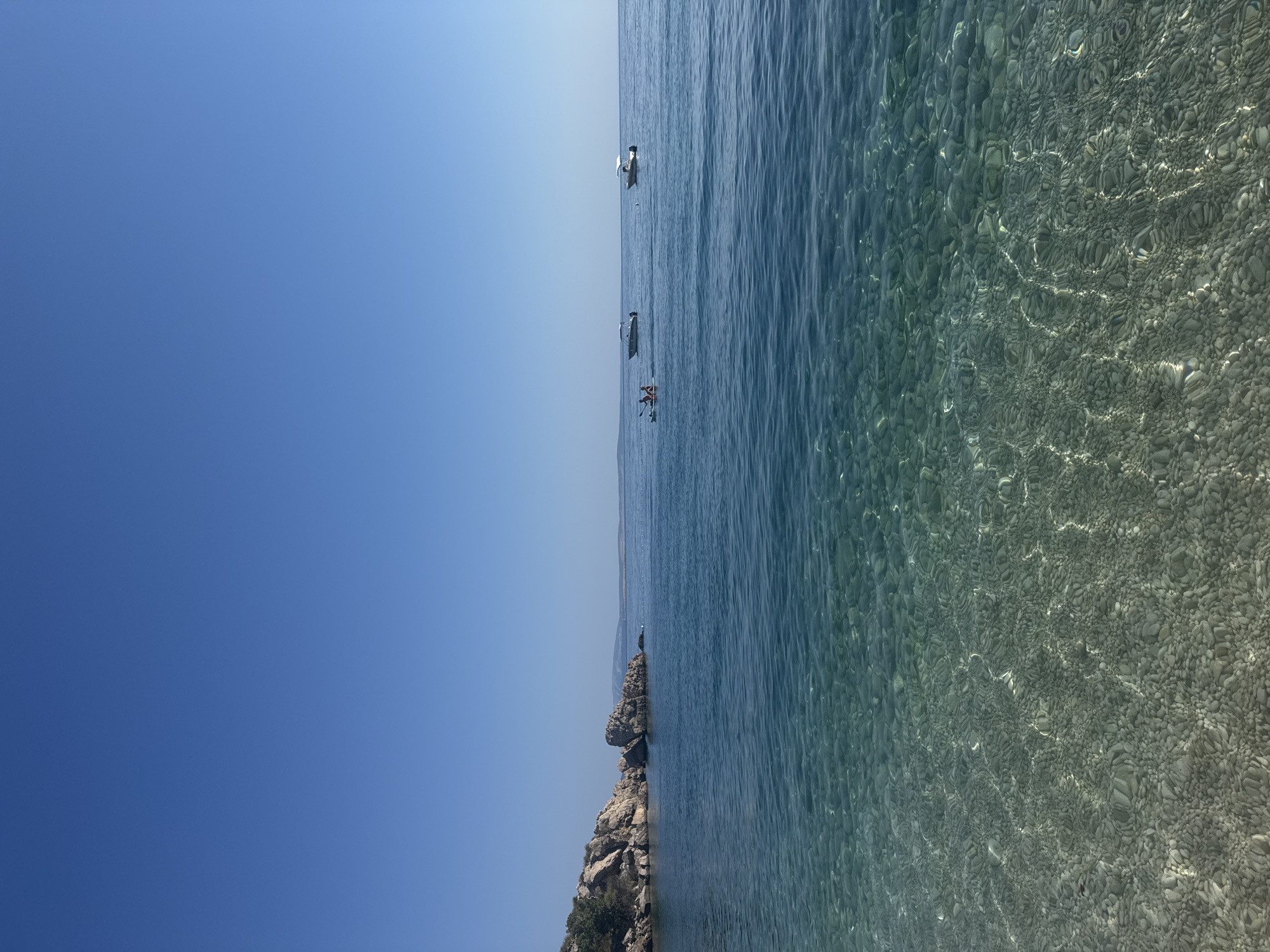 A bright, clear sky dominates the upper portion of the image, transitioning to a calm, blue sea that occupies the majority of the frame. The water is remarkably clear, revealing a seabed covered with smooth, rounded pebbles in shades of grey and white. In the middle ground, two small motorboats are anchored or moving slowly on the water. Closer to the left side, a kayak with two people is visible, appearing to be paddling. The left side of the image features a rocky coastline with sparse vegetation, characteristic of the Mediterranean landscape. The scene depicts a serene day at the beach in Stara Baška, Croatia.