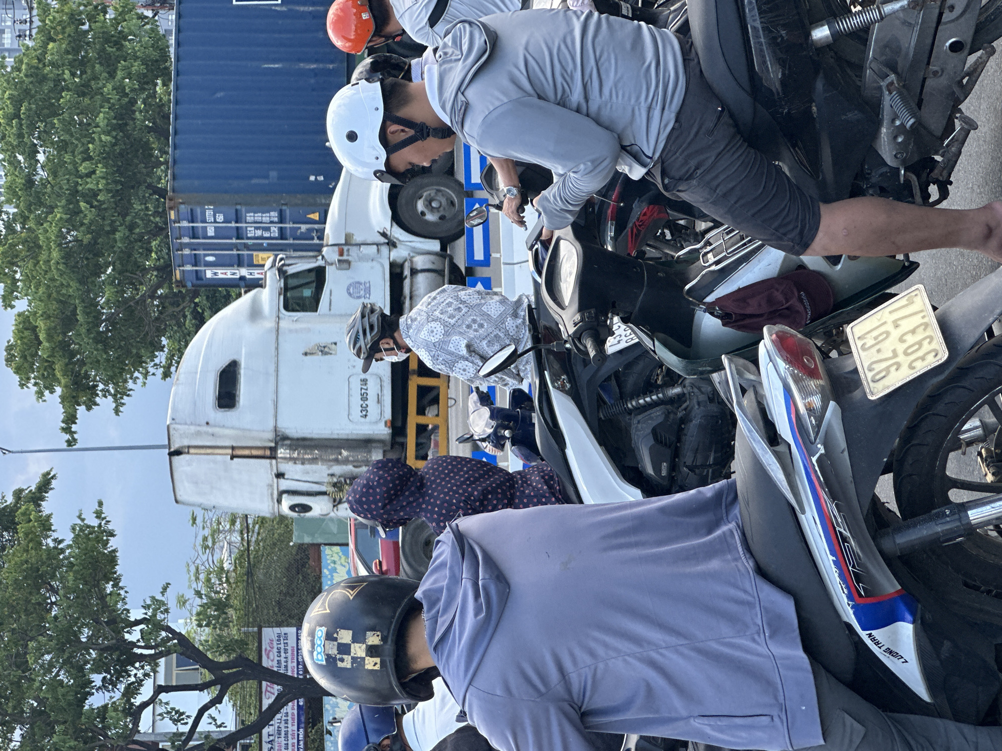 The image captures a street scene in Đà Nẵng, Vietnam, possibly during the daytime under clear skies, as suggested by the bright lighting. The primary focus is on several people on motorcycles, indicating a traffic or parking situation.
In the foreground, a person wearing a light blue hoodie and a black helmet with a checkered design is seated on a motorcycle. To their left, another person in a patterned shirt and a helmet is also on a motorcycle. Further back, a person in a grey hooded jacket and a white helmet is leaning over a motorcycle, possibly interacting with it or another person. An orange helmet is visible on another individual.
The background reveals a busy urban environment. A large white truck with the license plate "43C-05746" is prominently featured, suggesting it might be part of the scene's context, perhaps indicating a delivery or transport operation. A large blue shipping container is also visible. Trees and some buildings or signs are present, further defining the urban setting.
Several pieces of text are visible, though partially obscured or difficult to read entirely due to the image's orientation and resolution. Some legible text includes Vietnamese words like "SÁT T," "DAI THÉP LI," "GIA CÔNG V HỒ GA," and numbers like "8619-0935000 410" and "43C-05746." There is also text on a license plate which appears to be "19-76" and "393.74" and "860". Another motorcycle has text that reads "JUONG TRAN".
The overall impression is a snapshot of everyday life in a bustling Vietnamese city, with a focus on motorcycle traffic and the people who navigate it. There are no strong emotional cues visible; the scene appears to be a neutral observation of the environment and its inhabitants.