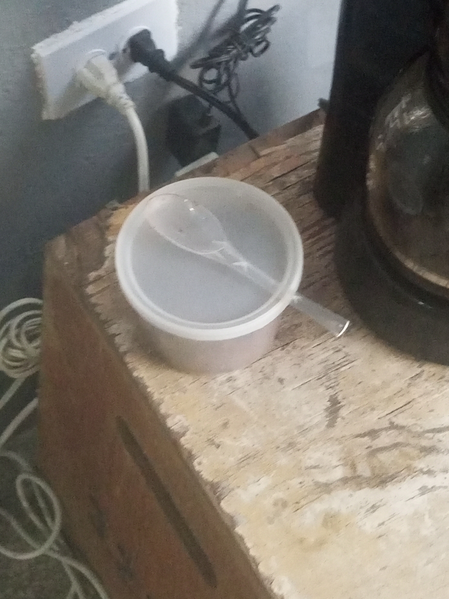 A close-up, eye-level shot captures a small, clear plastic container with a white rim and a translucent spoon resting inside it. The container sits on a weathered, light-colored wooden surface, likely a table or shelf. Behind the container, a white electrical outlet is visible, with a black electrical cord plugged into the right socket and a white cord plugged into the left socket. The white cord extends downwards, coiled partially on the left side of the frame. To the right of the container, a dark, cylindrical object with a black lid, possibly a coffee maker, is partially visible. The background is a textured, dark gray wall. The scene appears to be indoors, with artificial lighting. The location is identified as Juan Griego, Venezuela.