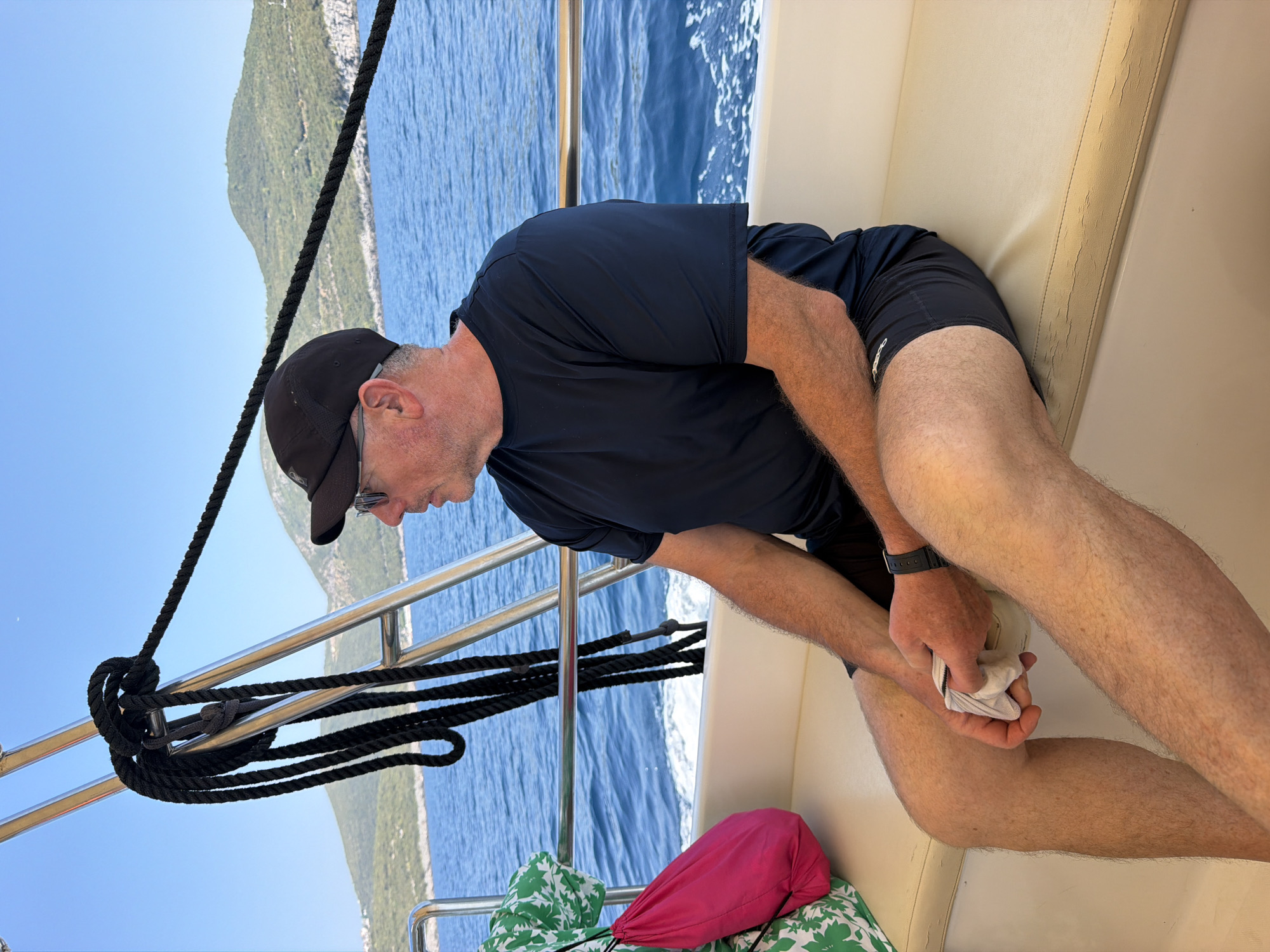 A middle-aged Caucasian man wearing a black baseball cap, sunglasses, a dark blue short-sleeved shirt, and black shorts sits on the cushioned bench of a boat. He is holding a white cloth in his hands. The boat is moving on the Adriatic Sea, with a wake visible behind it. In the background, green, hilly islands are visible under a clear blue sky. The scene is located in Dubraljevina, Montenegro. A pink bag and a green and white patterned item are to the man's left. A thick black rope is secured to the boat's railing.