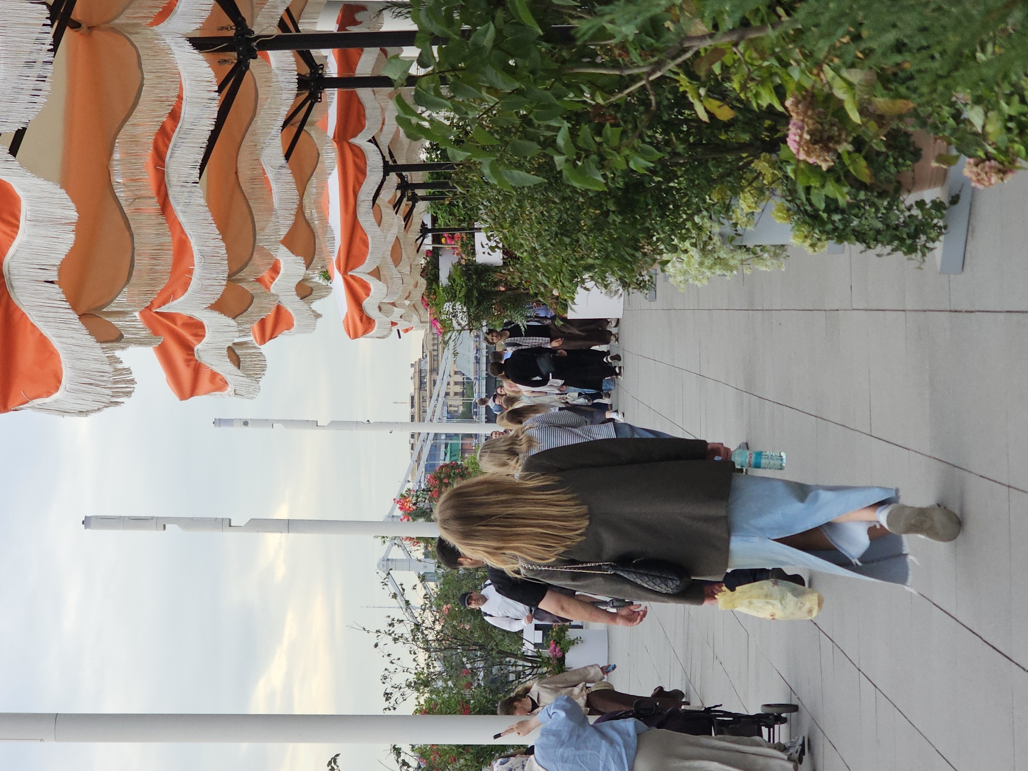 The image shows an outdoor scene with several people walking on a tiled walkway. Above them, there are large, decorative awnings with orange and white stripes, and white fringe. To the right, there are trees and plants, some with pink and white flowers. The sky is overcast, suggesting it is a cloudy day.
The people appear to be casually dressed and are walking in various directions. Some are walking away from the camera, while others are walking towards it or past it. One woman in the foreground is wearing a dark coat and a light blue skirt, and she is carrying a bag. Another person further back is wearing a white shirt and a black cap. The overall mood seems relaxed, as if people are enjoying a stroll in an outdoor setting, perhaps a park, a promenade, or a seating area at an event.
The scene suggests a daytime setting due to the brightness, but the overcast sky makes it difficult to determine the exact time of day. There are no visible signs or text that provide specific location information. The focus is on the people and the outdoor environment, with the colorful awnings and greenery adding visual interest.