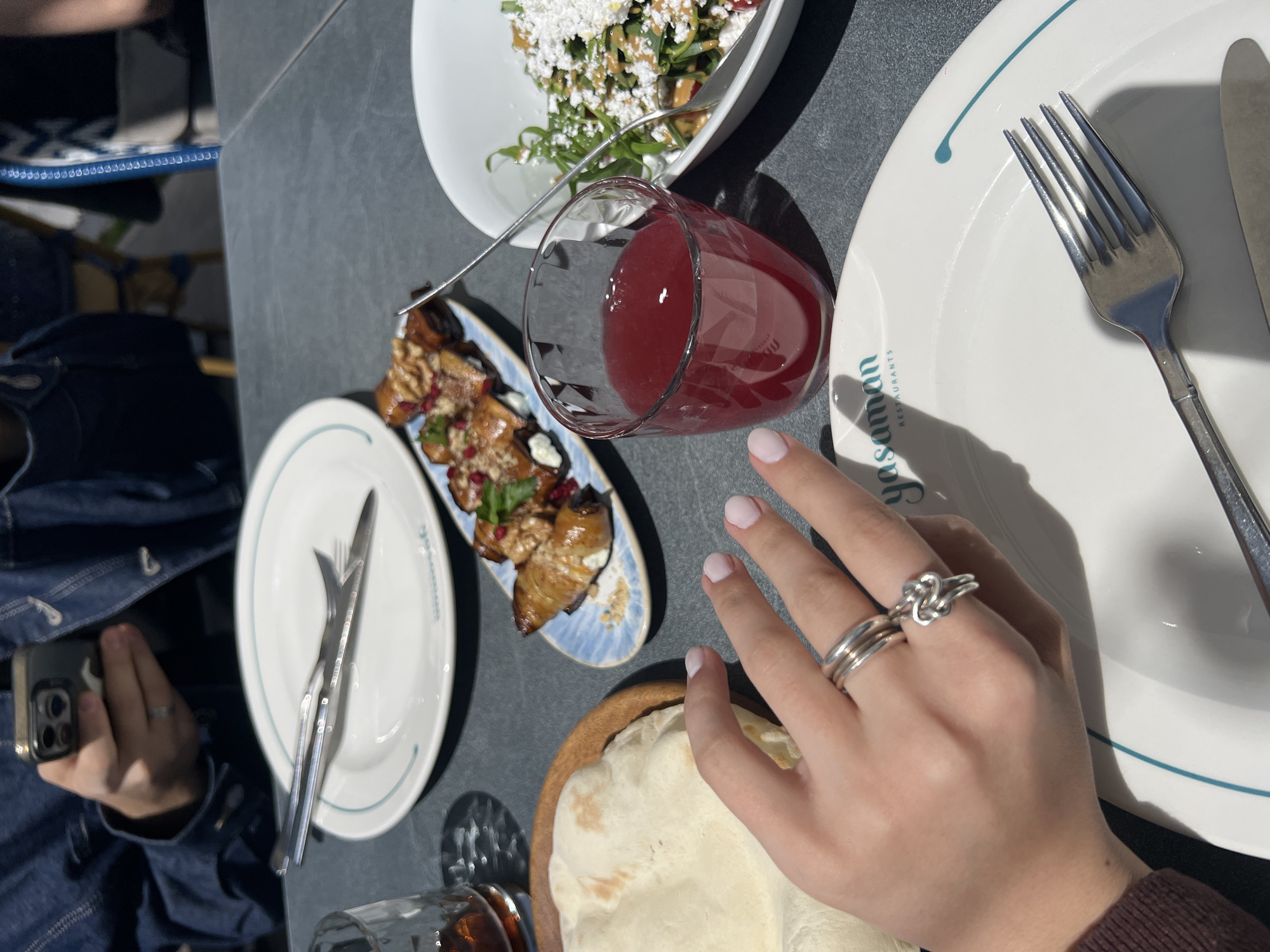 A close-up overhead shot captures a dining table scene, potentially at a restaurant called "Yasaman Restaurants." The table is a flat, gray surface, set with an assortment of dishes. A large white plate on the right side of the image contains a fork and knife, positioned neatly. Near the center, a glass filled with a red beverage is visible, along with a small plate holding a dish of eggplant. To the left, another white plate sits empty. A bread-like item in a wooden bowl is placed in the lower portion of the frame, adjacent to a hand adorned with several silver rings. A partial view of an individual holding a smartphone is also discernible in the bottom left. The location is described as Unknown, Unknown, indicating the city and state are unidentified. The scene suggests a shared meal or dining experience.