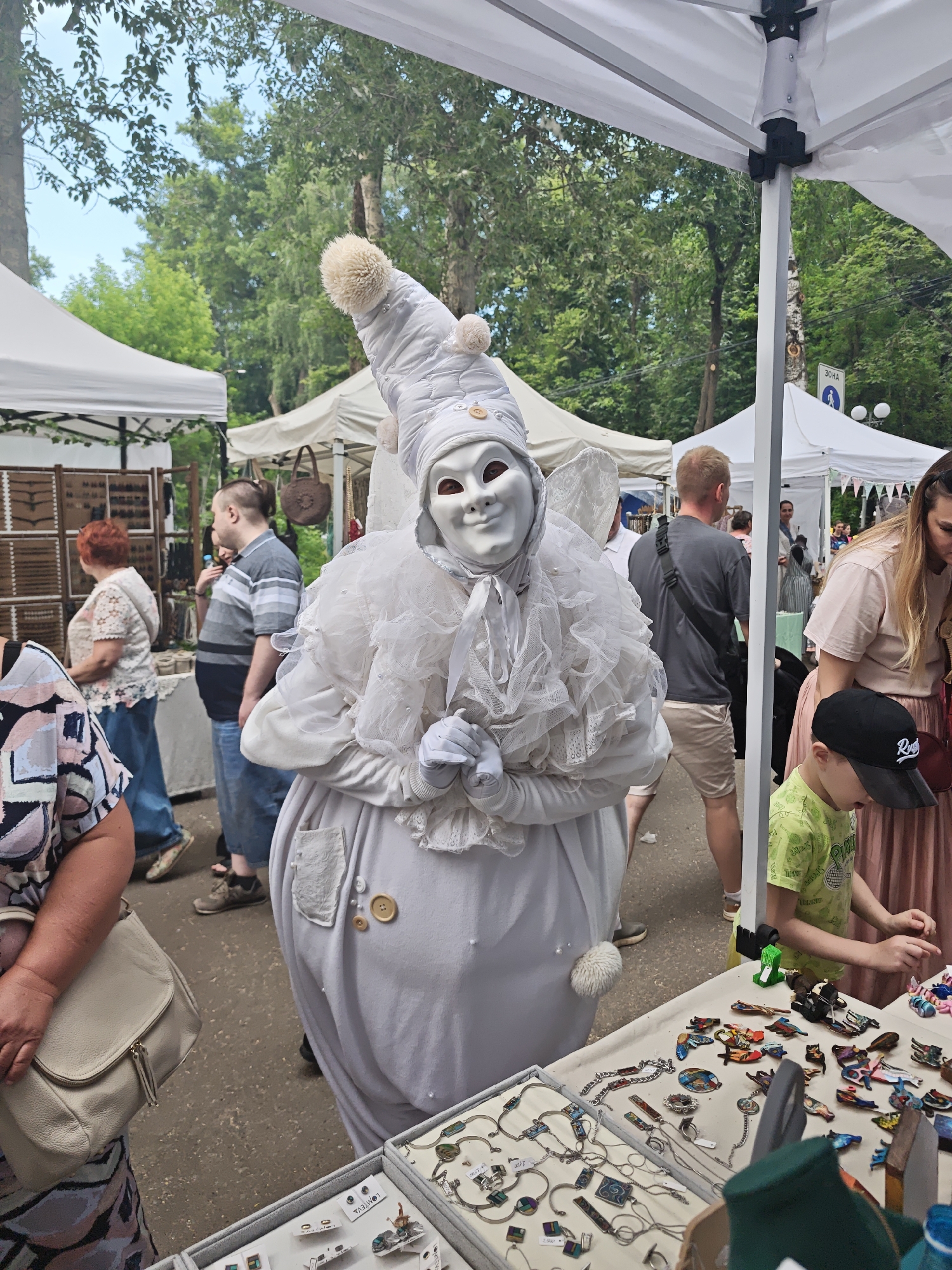In Kimry, Russia, an outdoor market scene unfolds under a bright sky. A performer in a white, elaborate costume, including a tall conical hat and a mask, stands prominently in the foreground. The costume features ruffled fabric around the neck and chest, and several buttons adorn the front. The performer wears white gloves.
To the left, a woman in a patterned shirt and jeans carries a light-colored purse. Behind her, a man in a striped shirt and jeans walks away from the camera. Further back, another woman with red hair is browsing a stall.
To the right, a table displays various small trinkets and jewelry, such as necklaces, brooches, and small figures. A young boy in a bright green t-shirt and a black baseball cap is leaning over the table, seemingly examining the items. A woman in a pink shirt and skirt stands behind him, partially visible. Tents and trees form the backdrop, suggesting a public gathering or festival in a park-like setting. The overall atmosphere appears to be lively and bustling.