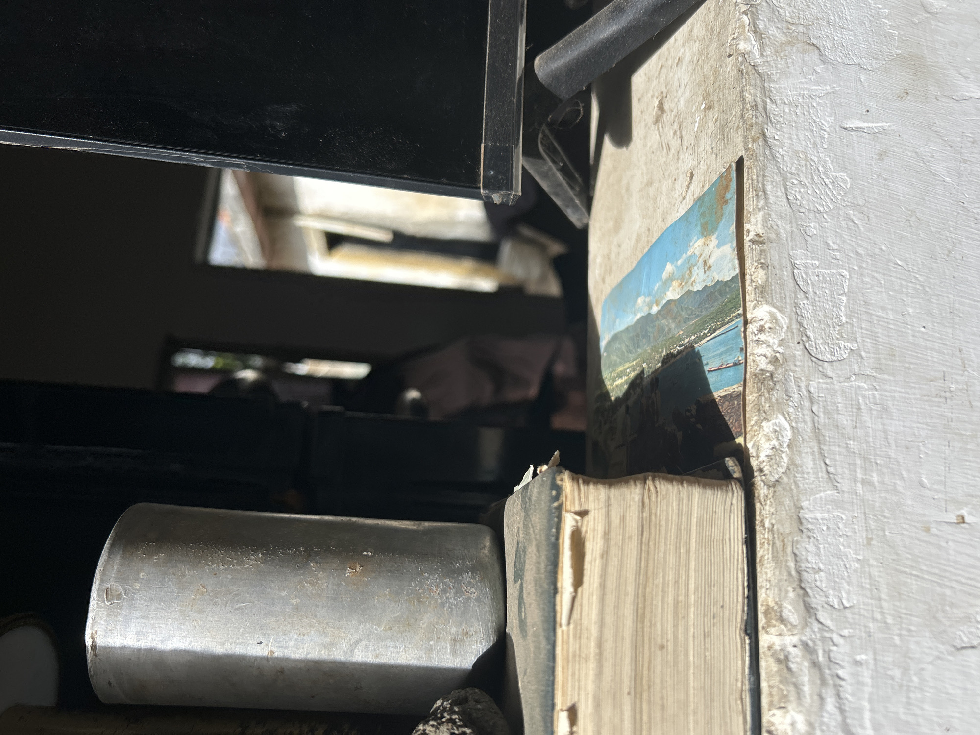 The image depicts a still life arrangement on a weathered, white ledge, possibly a windowsill or a table. In the foreground, a thick, aged book with yellowed pages rests on the ledge. A small, faded postcard, featuring a landscape with blue water and green hills, is placed on top of the book. To the left of the book, a cylindrical metal object, appearing to be a can or container, stands upright, casting a shadow. Behind these items, a black electronic device, possibly a monitor or television, is visible, propped up by a dark stand. The background shows a blurred view of what appears to be an outdoor setting, possibly a courtyard or street, with structures and natural elements. The overall scene is illuminated by strong sunlight, creating distinct shadows. The location is identified as Santa Ana, Venezuela.
