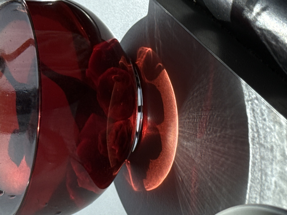 The image displays a close-up view of a clear glass, spherical bowl, located in Yerevan, Armenia. The bowl is filled with a dark red liquid and contains numerous dark red, irregularly shaped objects floating within it. The bowl rests on a flat, dark, reflective surface. A strong light source, positioned to the left, illuminates the scene. This illumination causes light to refract through the bowl and its contents, projecting a prominent, crescent-shaped area of bright red-orange light directly beneath the bowl's base on the underlying surface. From this central luminescence, intricate, radiating patterns of lighter and darker lines extend across the dark surface. The background immediately behind the bowl is uniformly light gray and out of focus. Portions of other indistinct objects are visible in the lower left and lower right corners of the frame. No people are present.