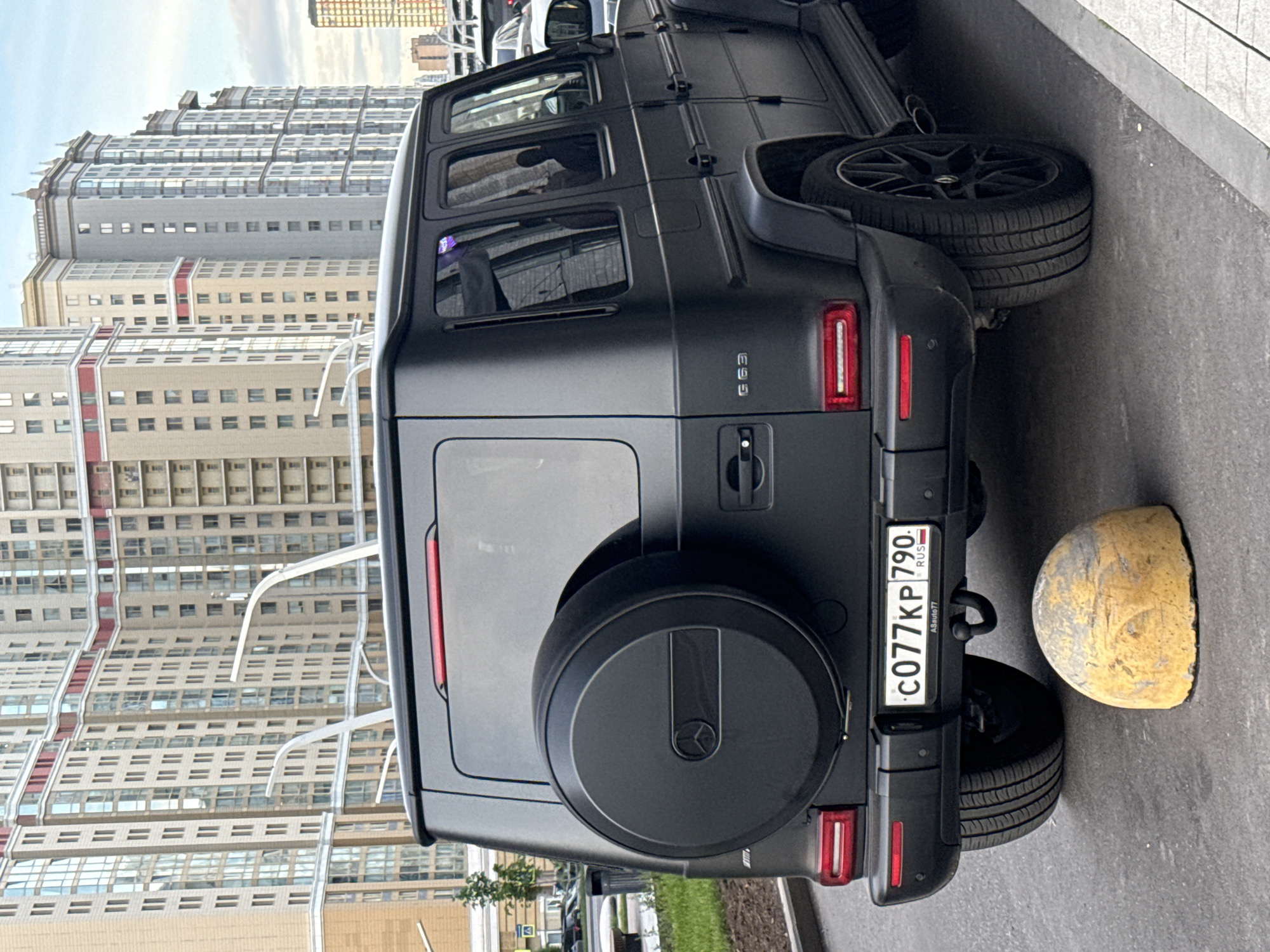 The image shows a matte black Mercedes-Benz G63 AMG parked near a sidewalk. The car has a spare tire carrier on the rear door. The Russian license plate is visible: C077KP 790. A yellow concrete bollard sits behind the car. The background shows a tall apartment building, which suggests a densely populated urban area. Based on the license plate and architectural style, the location is likely Moscow, Russia.