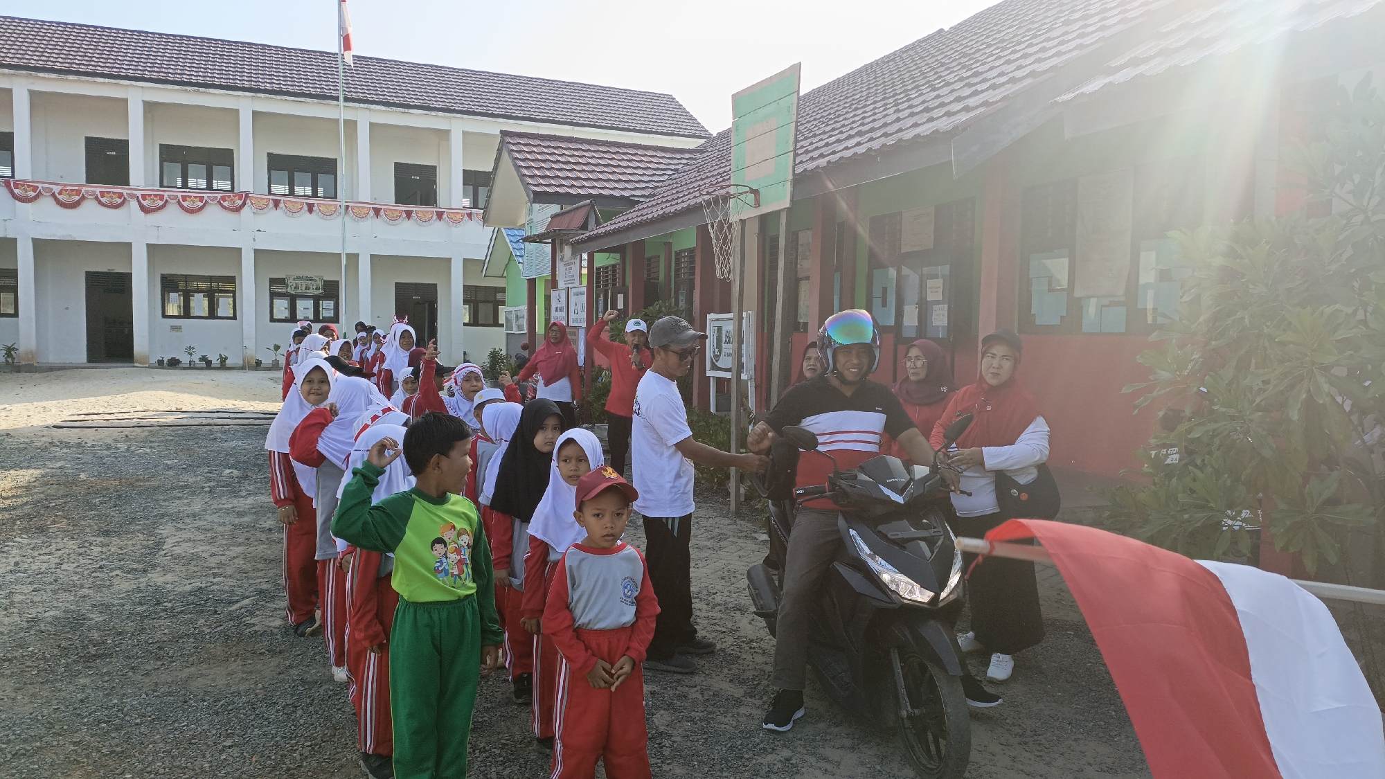 The image depicts an outdoor scene in Saring Sungai Binjai, Indonesia, likely at a school. In the foreground, a line of children wearing red and white uniforms and hijabs are assembled. Some children are looking forward, while others are engaged with adults. A young boy in a green shirt and green pants is prominent in the left foreground, looking to his right. To the right of the children, a man wearing a helmet and a black and white striped shirt is seated on a black motorcycle. He is interacting with an adult man standing next to him, who is wearing a white t-shirt and a grey cap. Several other adults, some wearing red headscarves, are visible in the background, observing the children or engaged in conversation. A large red and white flag, likely the Indonesian flag, is partially visible in the lower right corner, held by an unseen individual. Behind the children and adults, there are two buildings. The building on the left is a two-story white structure with red decorative trim and multiple windows. The building on the right is a single-story building with a red facade and a dark-colored roof, featuring a basketball hoop attached to its exterior. The ground is covered with gravel. The sun is shining brightly, casting lens flares and highlighting parts of the scene. The overall atmosphere appears to be that of a school event or assembly.
