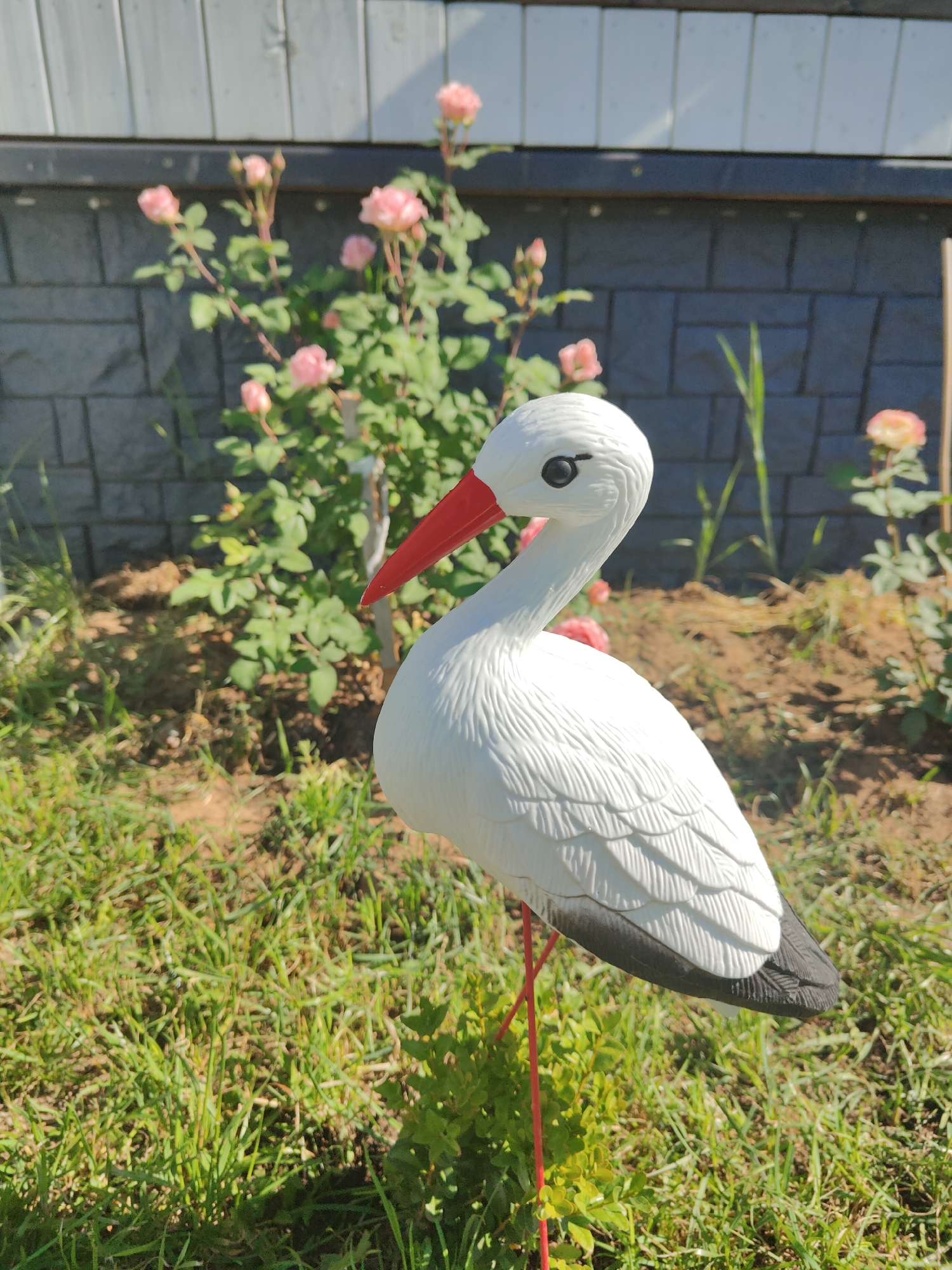 In Ozyory, Russia, a garden features a white plastic stork figurine standing on thin red legs. The figurine has a red beak, black wingtips, and a black eye. It is positioned in front of a rose bush with pink flowers and a grey brick wall topped by a light-colored horizontal fence. The ground is covered in green grass and brown soil.