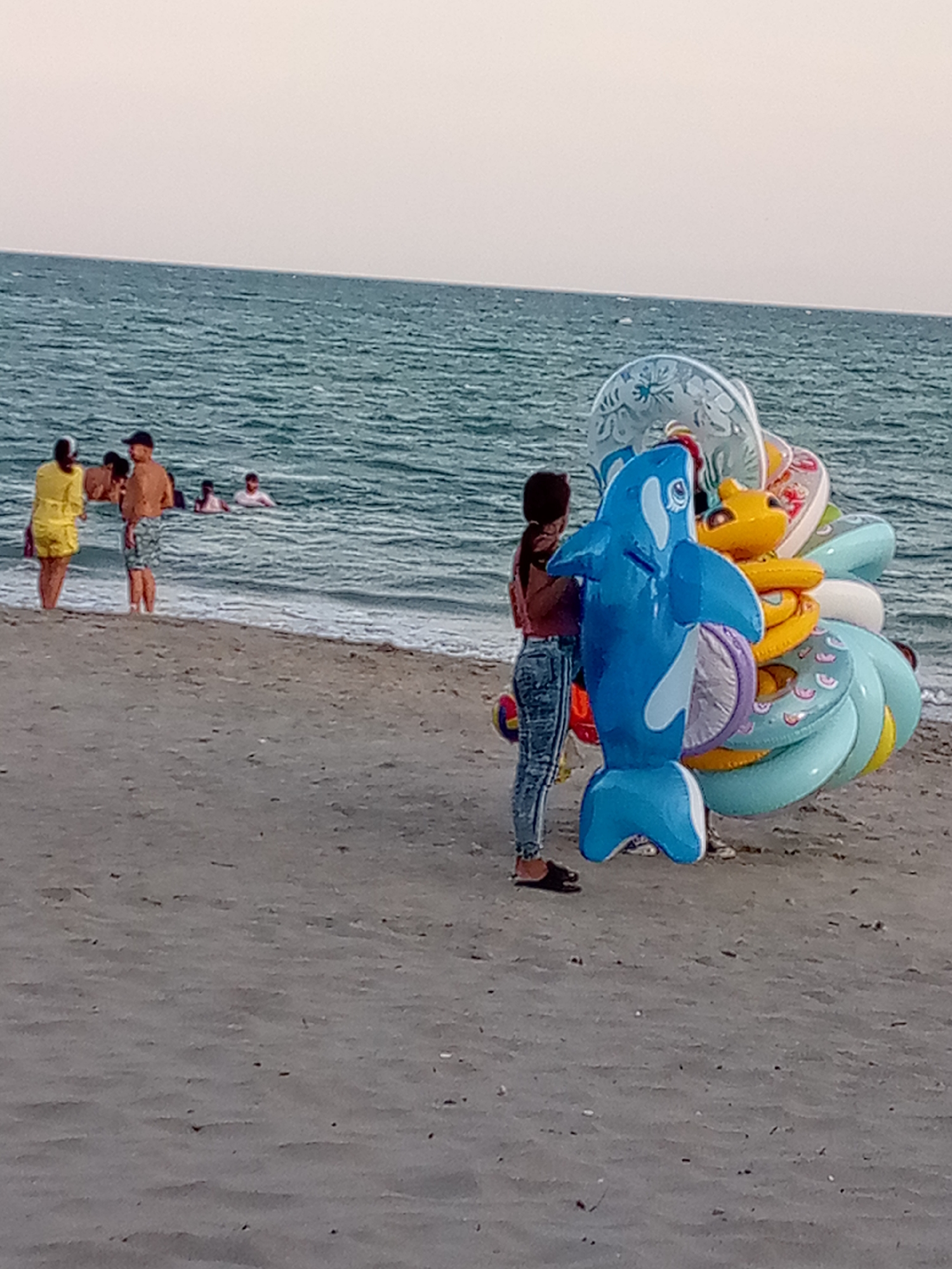 The image shows a beach scene in El Yaque, Venezuela. The foreground is dominated by a sandy beach, and a young woman stands near a large stack of inflatable toys. The toys consist of colorful swimming rings and a large, bright blue inflatable orca. The woman wears a pink top and jeans. Behind her, the ocean stretches to the horizon under a pale sky. Several people are visible at the water's edge, some standing and others swimming.