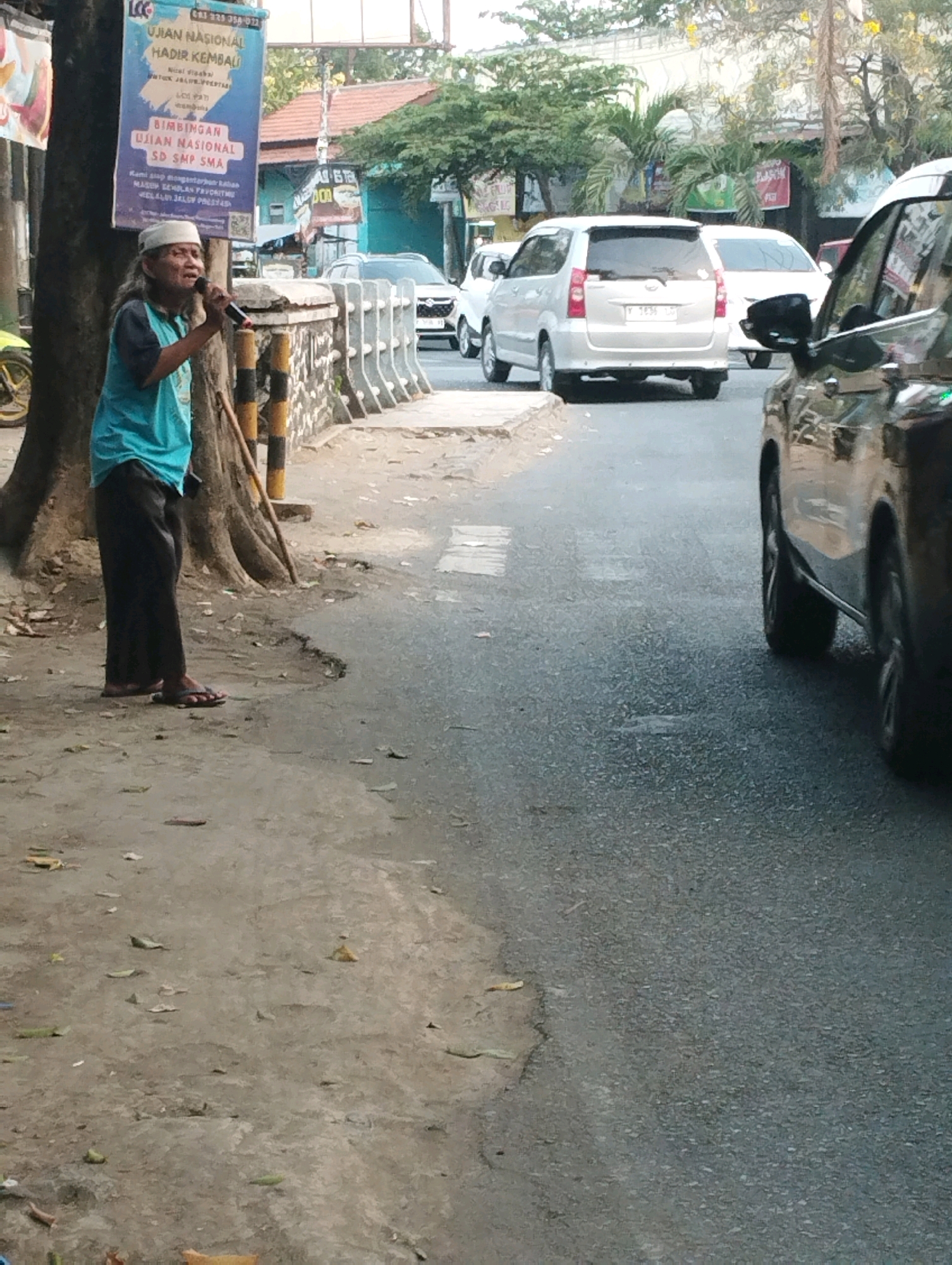 A man with long, gray hair and a white headscarf stands on a dirt path next to a tree, holding a microphone to his mouth. He is wearing a teal t-shirt and dark pants. A wooden stick leans against the tree. Behind him, a sign is attached to the tree trunk, with text in Indonesian. A white wall with black and yellow striped posts runs along the edge of the path. In the background, cars are driving on a paved road, including a silver SUV and a black car. Buildings and trees are visible further in the background. The scene appears to be in Kecamatan Pati, Indonesia, based on the sign's content.
