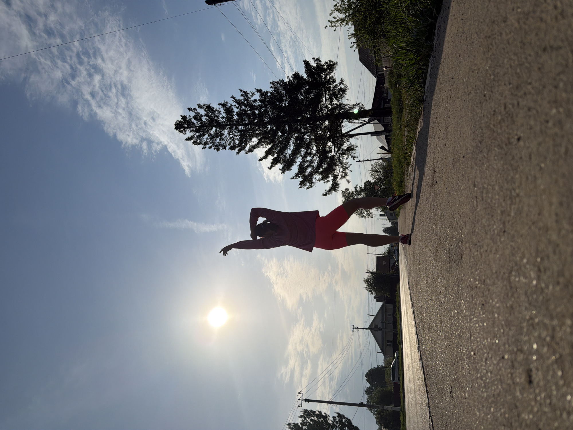 A low-angle shot, possibly taken with a mobile phone, presents a street scene in what appears to be a residential area, likely in a city. The photograph is tilted approximately 90 degrees counter-clockwise. The lower portion of the frame shows the sun prominently in the sky, which is partly cloudy. In the center, a person is captured in motion, seemingly stretching, wearing athletic clothing: a pink top and red shorts. The person's silhouette is cast against the bright sky. The background includes power lines, trees, and the roofs of houses, giving context to the scene. The foreground displays a pavement and street, suggesting the person is exercising or walking on a side street. Considering the context provided, it is likely this photograph was taken in Chaikovsky, Russia.
