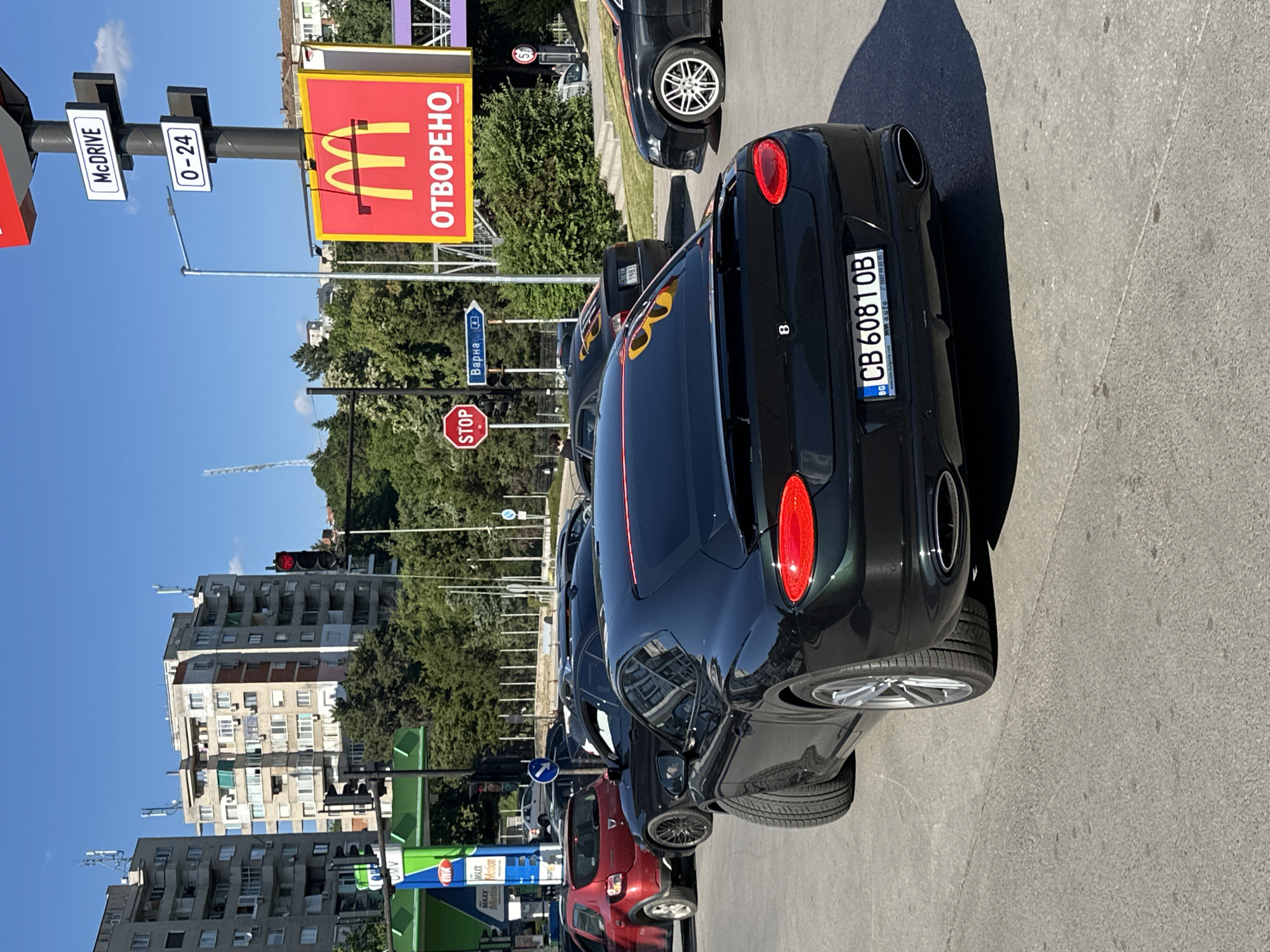 This is a vertical, eye-level, outdoor photograph taken on a sunny day, likely in the afternoon, in Veliko Tarnovo, Bulgaria. The scene depicts a street with several cars lined up, suggesting traffic or a queue. Dominating the foreground is the rear of a black sports car. The car's license plate is visible, reading "CB 60810B".
To the left of the cars, a tall pole displays signs for a McDonald's restaurant, with "McDRIVE" and "0-24" indicating it's a 24-hour drive-through. A larger McDonald's sign with the iconic "M" logo is also prominently featured, along with the Bulgarian word "ОТВОРЕНО", meaning "OPEN". Below this, a blue sign points towards "Варна", a city in Bulgaria. A red circular "STOP" sign is also visible.
In the background, there are multi-story apartment buildings with numerous windows and balconies, typical of Eastern European architecture. Green trees line the street, providing a contrast to the urban environment. The sky is clear and blue, suggesting fair weather. The overall impression is a snapshot of everyday urban life in a Bulgarian city, with a focus on the McDonald's branding and the cars present. There are no visible people in the frame.