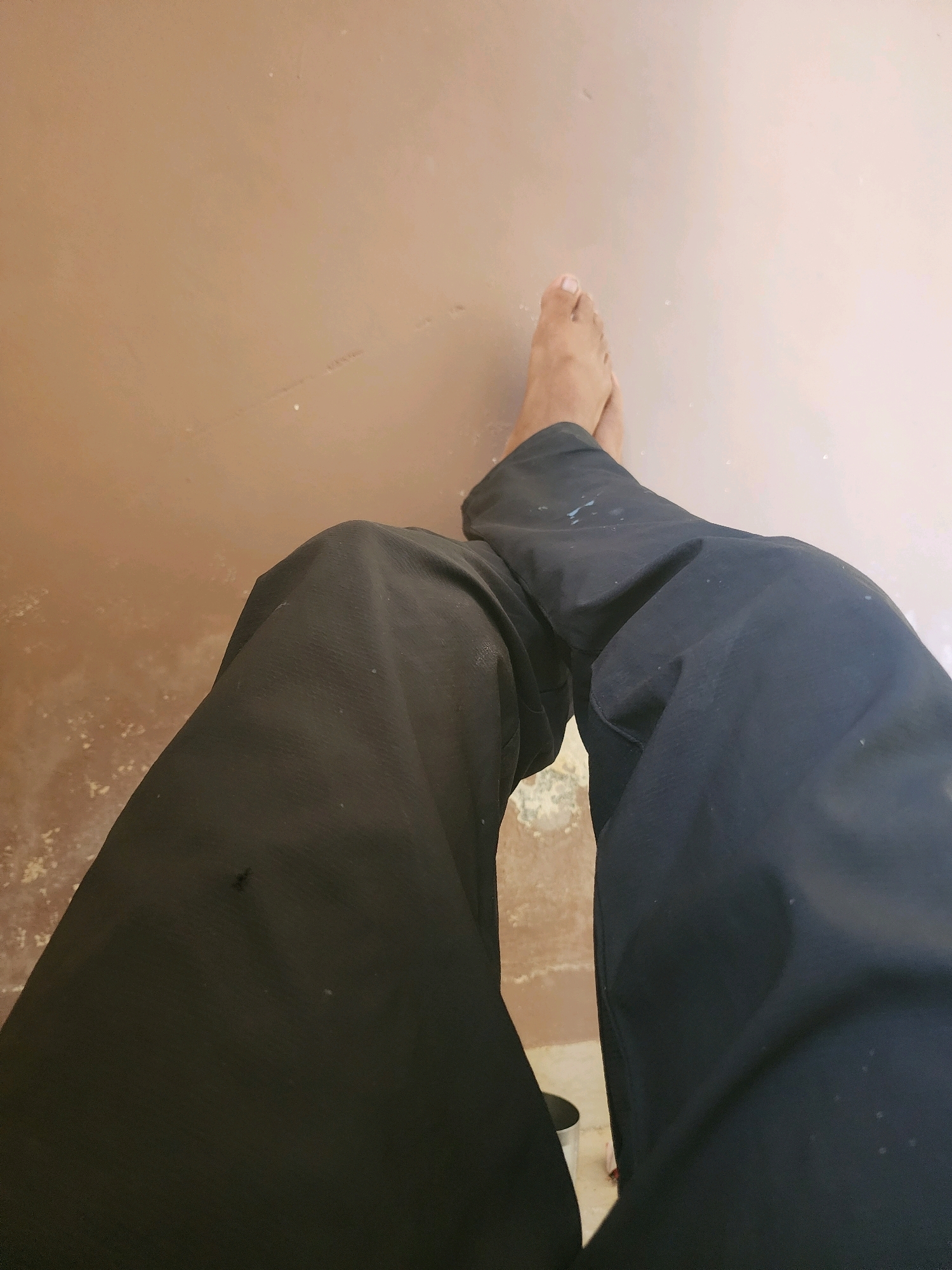The image shows a close-up view of a person's legs wearing dark-colored pants. One foot, bare and with visible toes, is visible at the top right of the frame, resting on a light brown, textured surface. The pants are loosely fitted and appear to be made of a fabric with some texture. There are small white specks on the fabric, possibly paint or dust. The lighting is even, suggesting an indoor or shaded outdoor environment. The background is a plain, light brown surface with some subtle variations in color and texture, appearing to be a floor or wall.