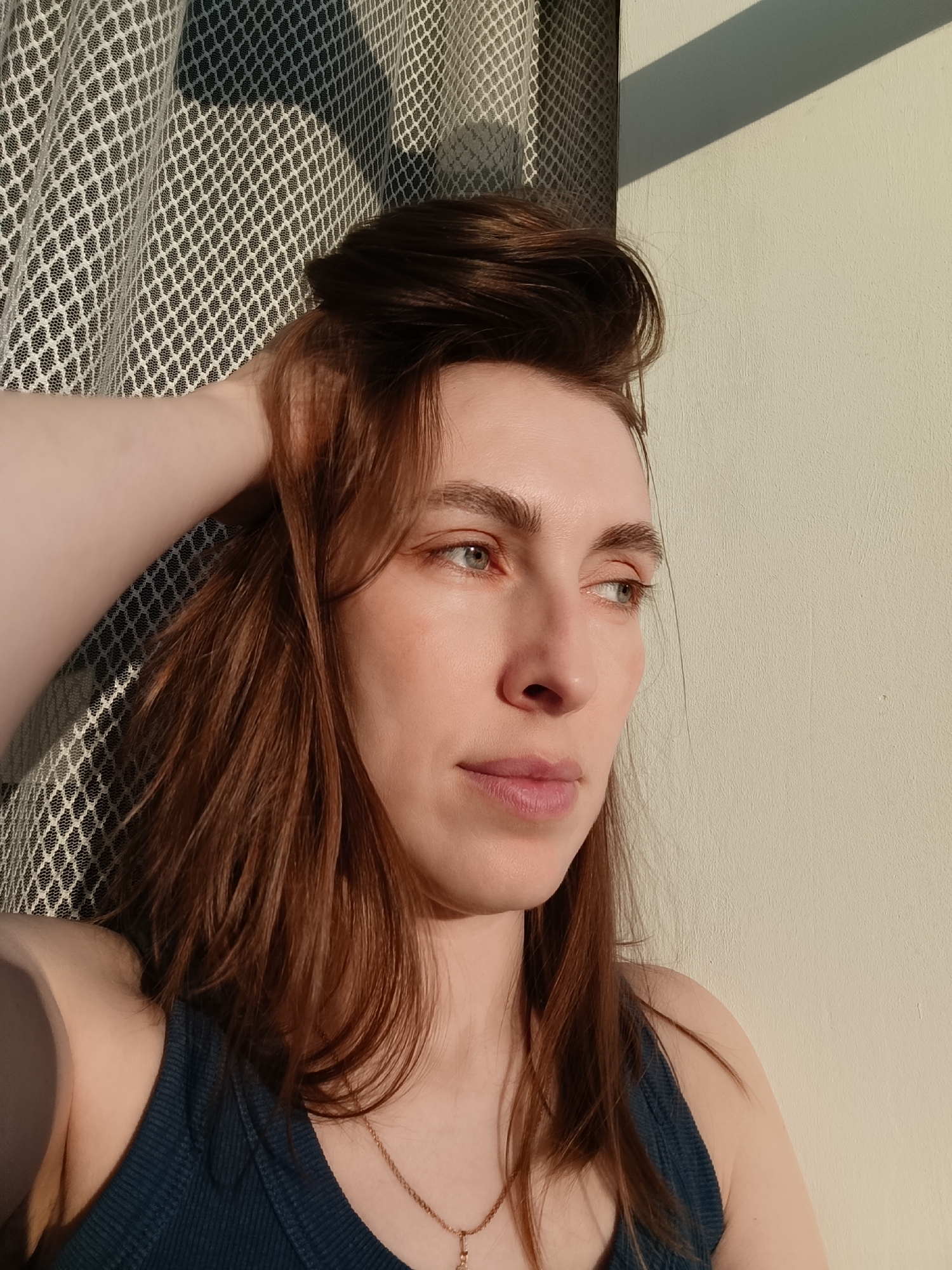 A fair-skinned woman with auburn hair is the subject of this portrait-style photograph. Her hair is partially styled in a bun at the crown of her head, with some strands falling around her face and shoulders. She is wearing a dark blue tank top and a delicate gold necklace. Her right hand is raised, with her fingers running through her hair. Her facial expression appears thoughtful, as she gazes off to the right of the frame. The light is predominantly warm, casting shadows on the right side of her face and body, indicating a possible setting sun or golden hour. The background consists of a wall, cream or off-white in color, and a sheer curtain with a diamond-shaped pattern. The city of Chaikovsky, Russia is not mentioned in the prompt. Therefore, it is not possible to provide a location.