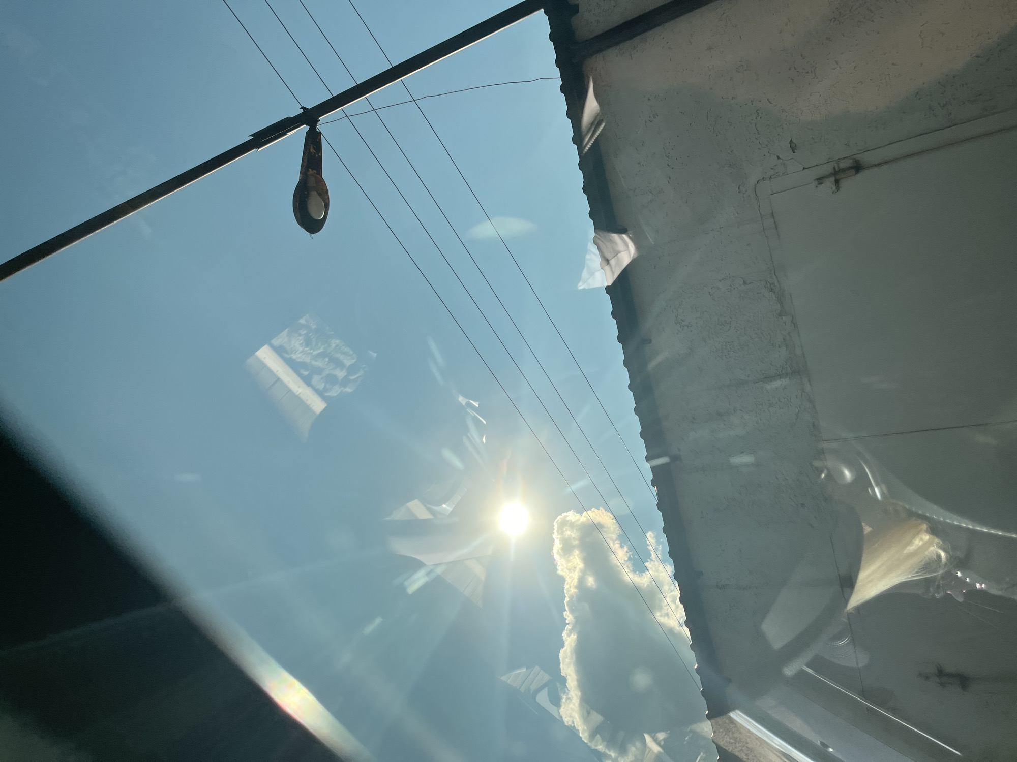 The image, captured through a glass surface, displays a bright sun and a clear blue sky. In the foreground, reflections on the glass obscure parts of the scene, including what appears to be a person's blonde hair and the interior of a vehicle. A large, fluffy white cloud is visible in the sky, with the sun casting bright rays. A street lamp with a rusty casing is mounted on a pole, with several power lines running horizontally across the sky. Below the sky, a textured, light-colored wall with a corrugated metal roof is visible, and a metal door with a latch is present on the wall. The overall scene suggests an outdoor environment, possibly a street or yard in an urban setting. The metadata indicates the location as Saratov, Russia.
