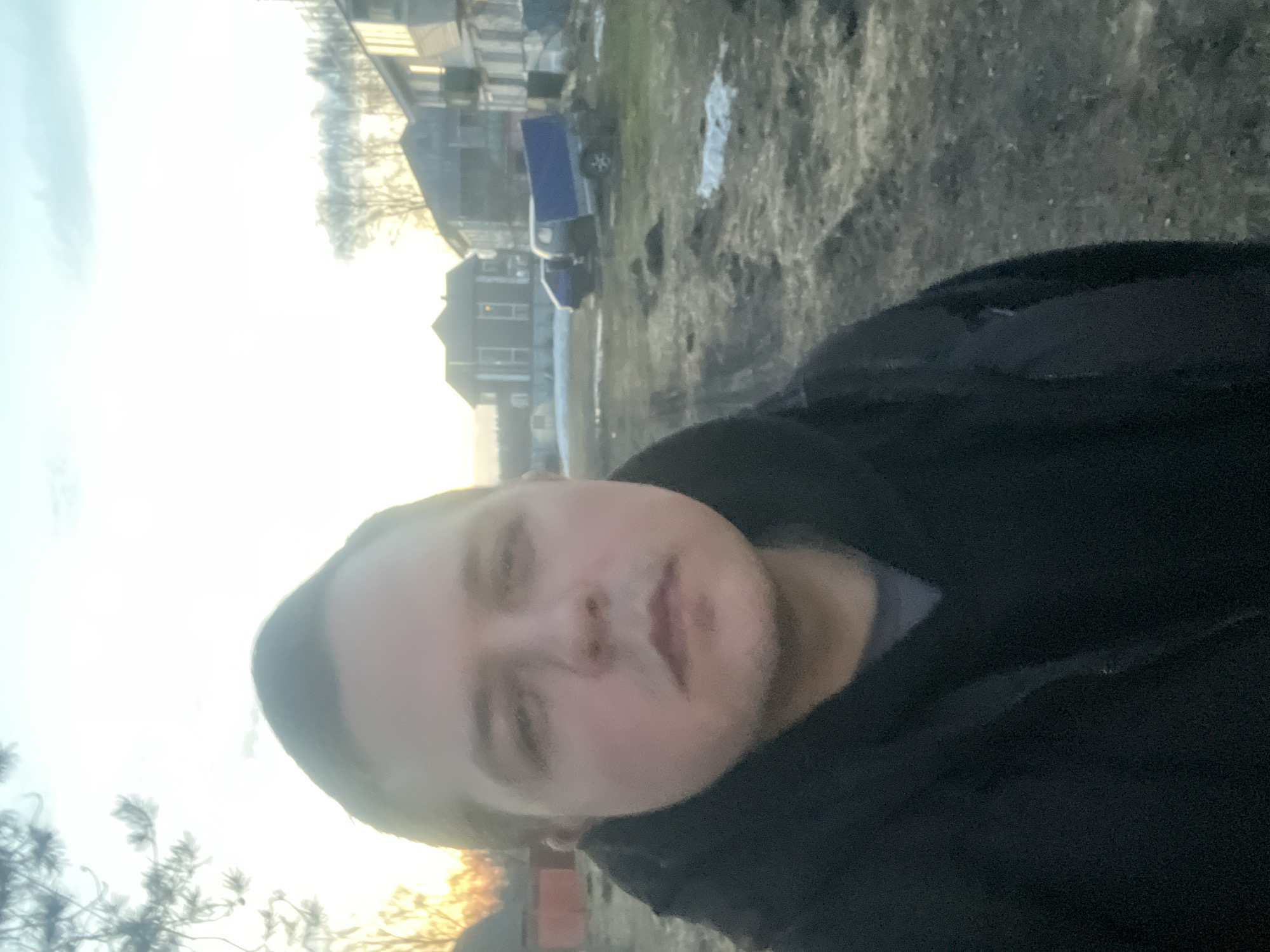 A medium, low-angle shot presents a person facing the camera, with residential buildings visible in the background. A light-colored sky dominates the upper portion of the frame. The ground appears to be a mixture of dirt and sparse vegetation. In the middle ground, buildings are visible, with what appears to be a blue truck parked nearby. Based on the location, the scene is set in Раздолье, Russia.