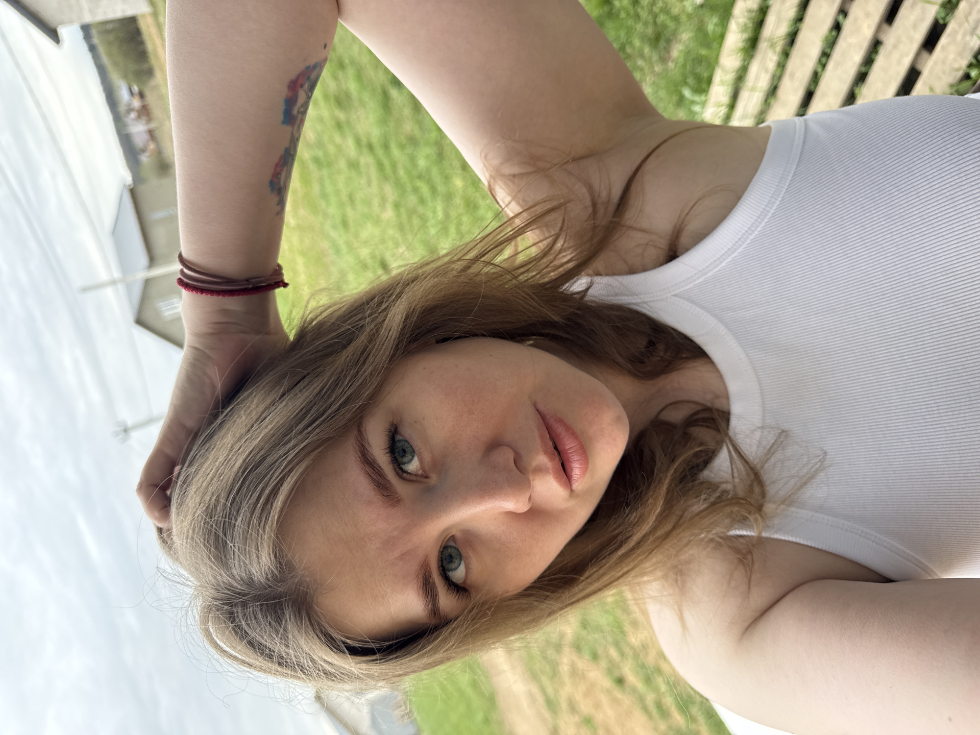 A young woman with light brown hair and blue eyes is looking at the camera. She is wearing a white tank top and has a tattoo on her left arm. The background shows a green lawn, a fence, and a building. The photo was taken in Ферма, Russia.