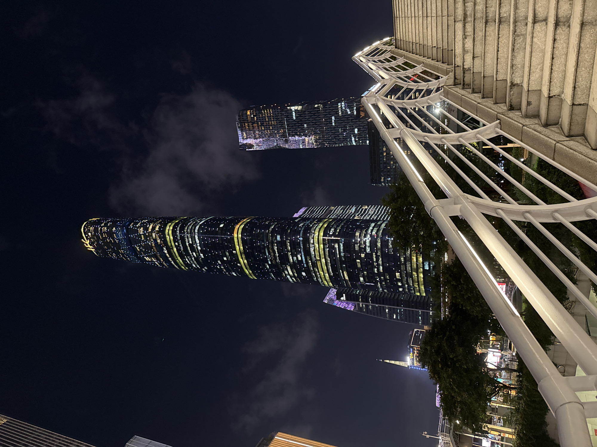 The image shows a night cityscape in the Tianhe District, China, featuring illuminated skyscrapers under a cloudy sky. The buildings are adorned with multiple lights that create a unique visual effect. In the foreground, there is a white architectural structure composed of bars and a stone staircase on the right.