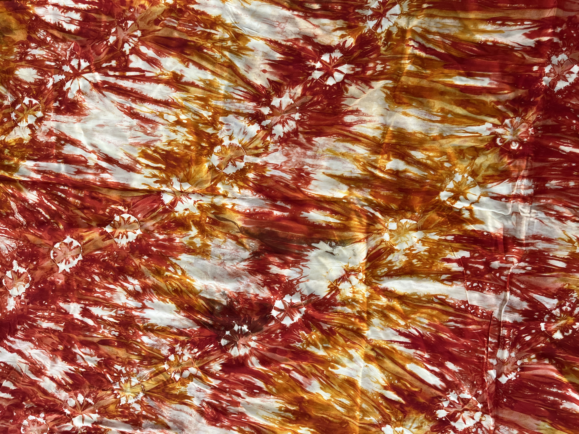 The image shows a close-up of a piece of tie-dye fabric. The fabric is predominantly white with red and orange dyes creating a radial pattern. The exact location in Jos, Nigeria is not discernible from the image. There are no people, specific objects, actions, interactions, or details about the environment visible.