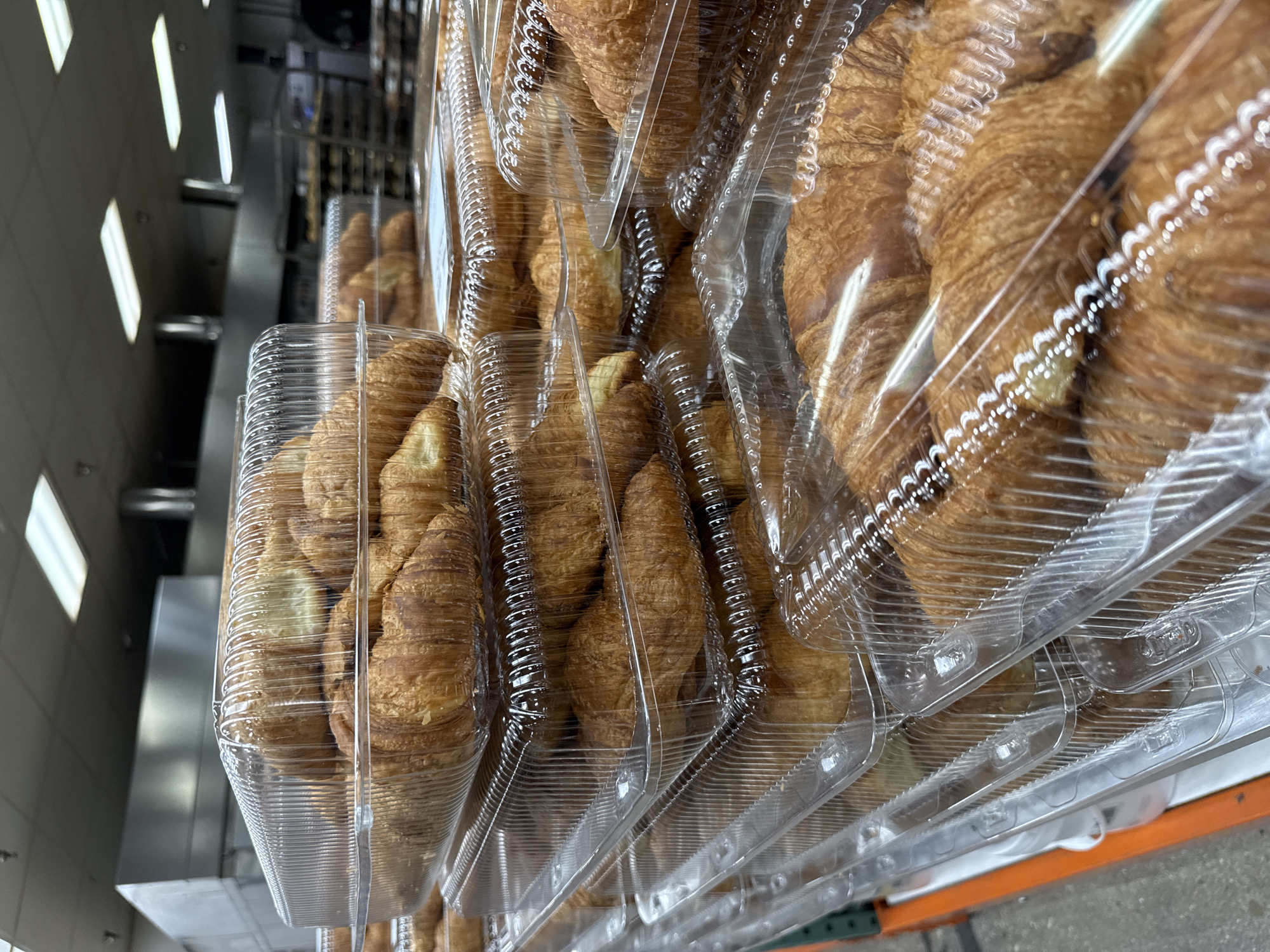The image shows a stack of clear plastic containers filled with croissants. The containers appear to be stacked on a shelf or display, possibly in a store setting, considering the fluorescent lighting visible in the background. The croissants are golden brown and appear freshly baked. The setting is likely a Costco store located in Davie, Florida, United States, based on the common practice of Costco selling croissants in such large, clear plastic containers.