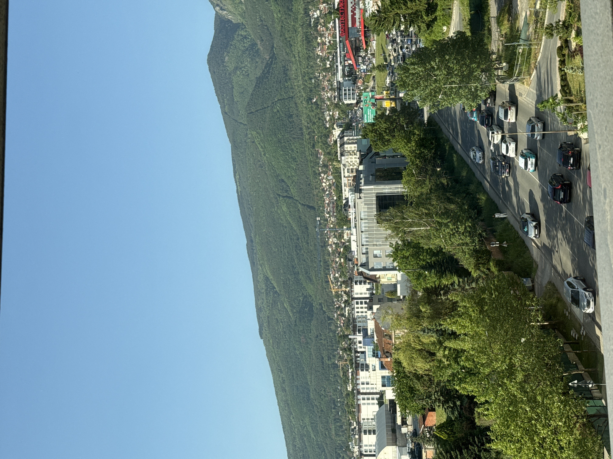 The image depicts a view in Sofia, Bulgaria, featuring a mountain range densely covered in trees to the left, under a clear blue sky. To the right of the mountains are buildings, including high-rise apartments and commercial structures, densely packed together. A main road runs parallel to the buildings, with cars traveling along it in both directions. There is greenery, including trees and shrubs, interspersed among the buildings and along the road. A cable car system with red supports and multiple cabins is visible toward the center, near the top of the buildings. The scene is taken from a high vantage point, possibly a balcony or rooftop.