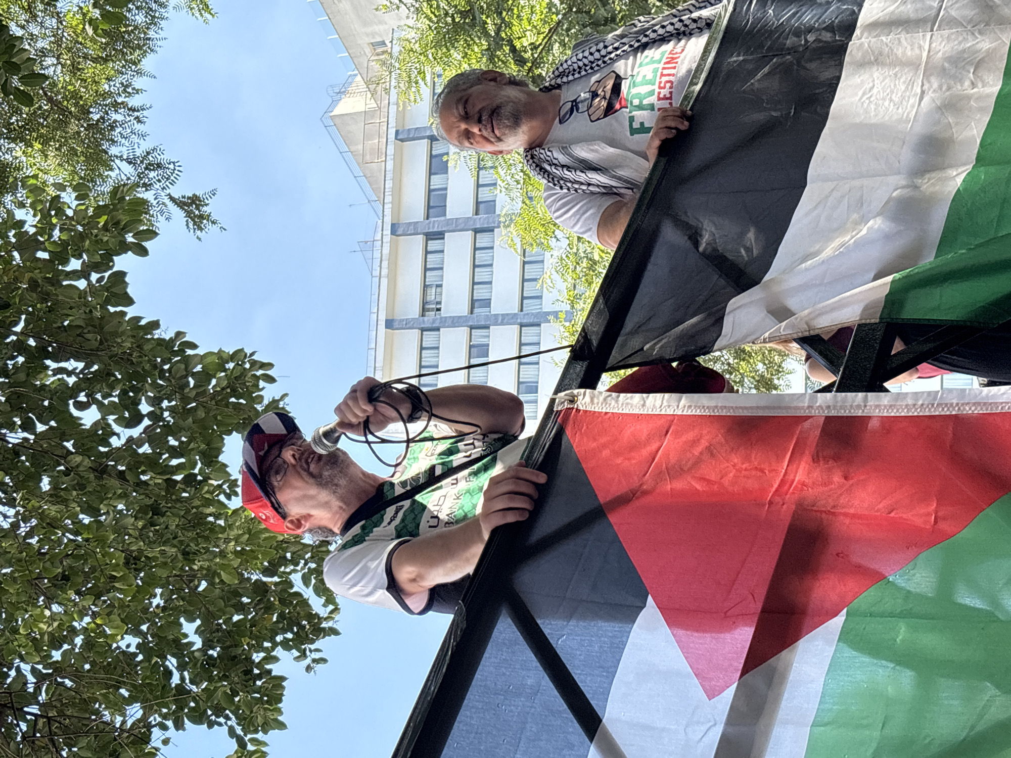 A low-angle, medium shot captures two men and a partially-visible flag against a backdrop of trees, a blue sky, and a tall building. The man in the foreground is speaking into a microphone held in his right hand. He has a fair complexion, a short beard, and wears a red and white hat, glasses, and a white t-shirt with a green design. The second man, positioned behind and to the right, is looking downward and smiling. He has fair skin, a gray beard, and wears a keffiyeh around his neck and a white t-shirt with the words "FREE PALESTINE" printed on it. They are holding onto the edges of a large flag featuring the colors black, white, green, and red. The scene appears to be outdoors, with trees visible on the left side. The presence of the tall building suggests an urban environment, specifically the city of São Paulo, Brazil.