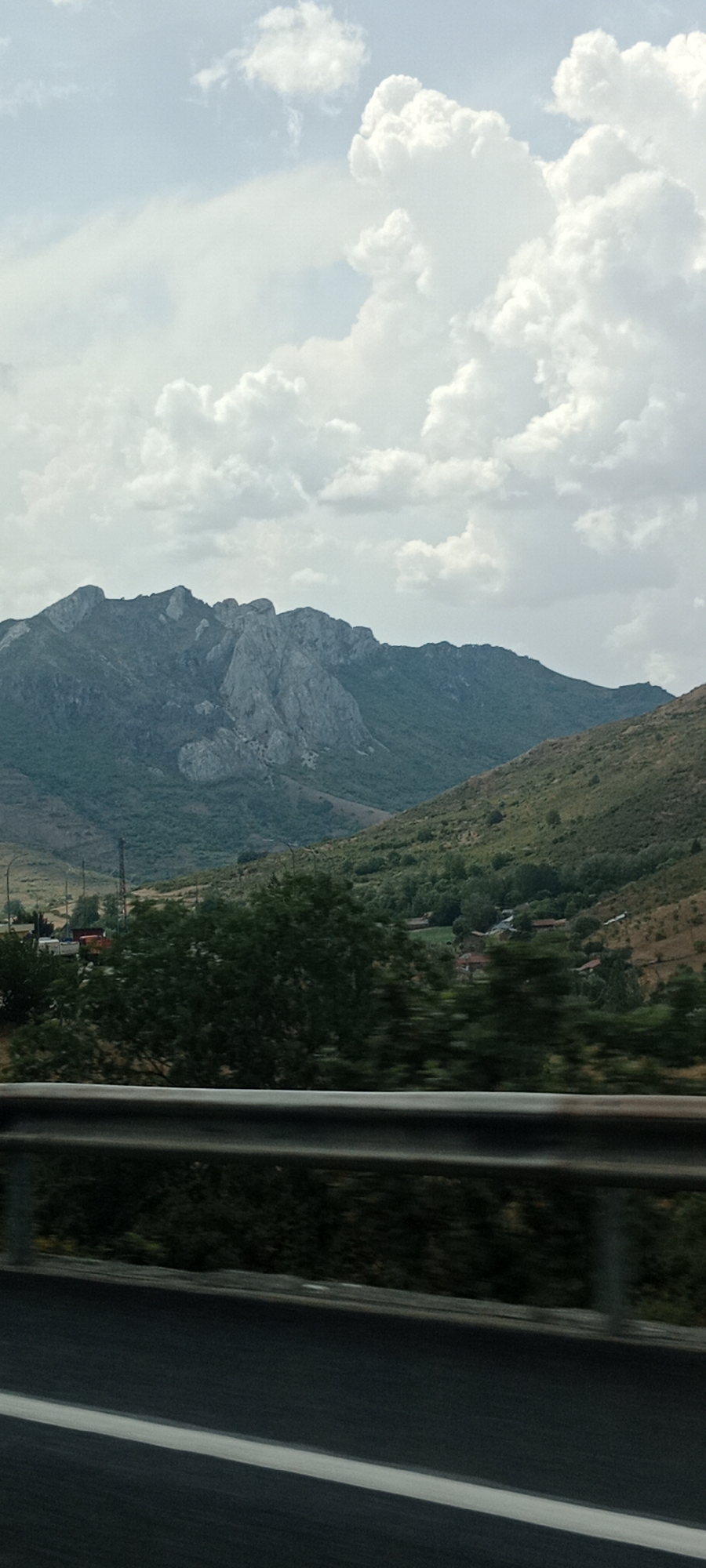 A vertical, slightly blurry, outdoor photograph taken from a moving vehicle captures a mountainous landscape under a cloudy sky. In the foreground, a dark asphalt road with a white lane marker runs horizontally across the bottom of the frame, bordered by a silver metal guardrail. Beyond the guardrail, a dark, blurred expanse of vegetation is visible, suggesting a roadside embankment or trees.
The mid-ground of the image displays a verdant valley dotted with small buildings, some with red roofs, nestled among lush green trees. The valley floor slopes upwards into rolling hills covered in vegetation and scattered trees. In the background, a large, rugged mountain range dominates the scene. The mountains are characterized by steep, rocky slopes with significant rock formations, their peaks jagged and irregular against the sky. The overall impression is of a rural, mountainous region. The location is identified as Sena de Luna, Spain.