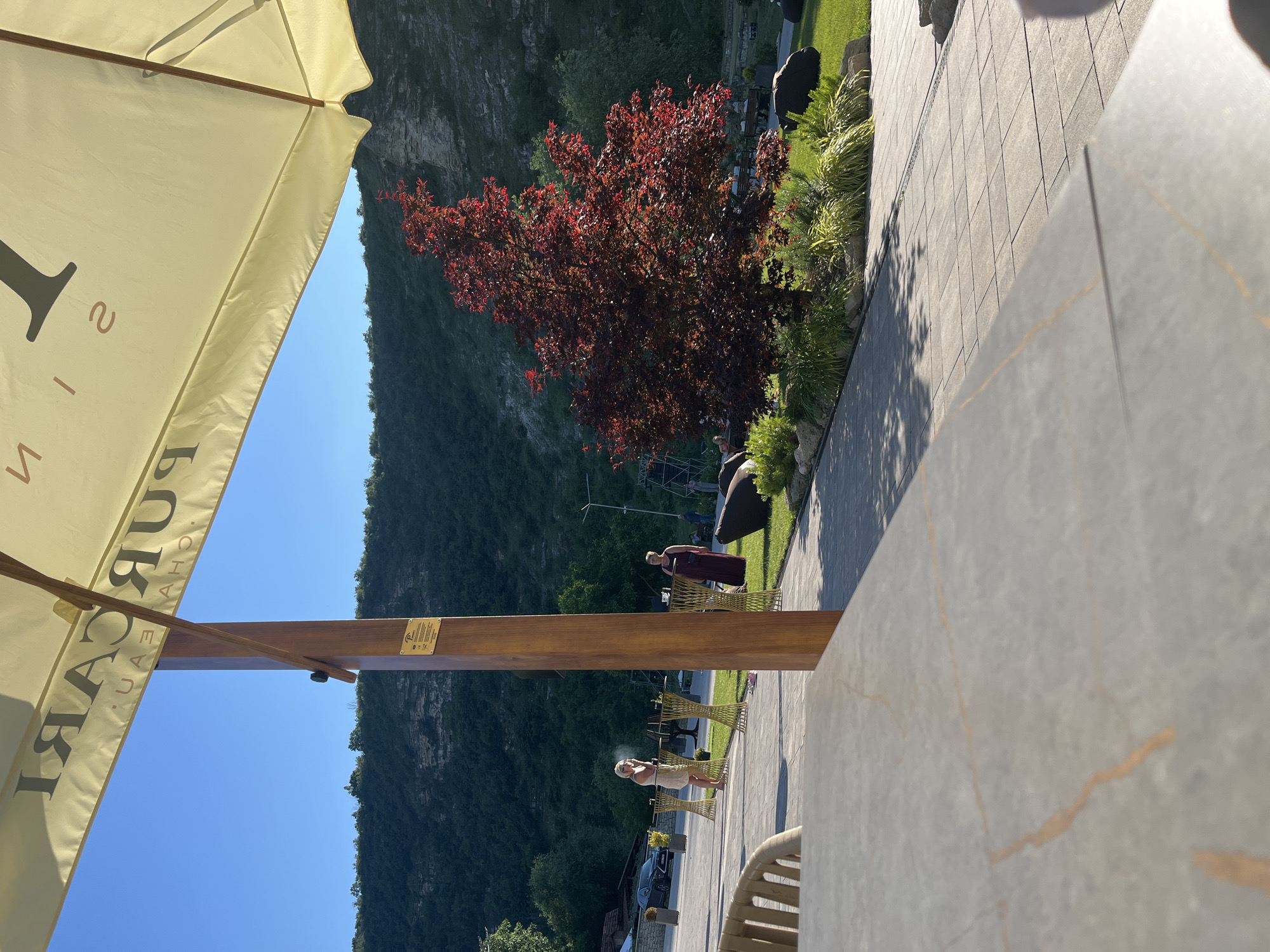 The image captures a sunny outdoor scene at Chateau Purcari, a winery in Butuceni, Moldova. In the foreground, a portion of a light-colored umbrella with text in black is visible, casting a shadow. A wooden pillar supporting the umbrella is also present. To the left, a woman in a light-colored dress stands near some gold-colored decorative stands. Further into the scene, a woman in a dark dress stands near a green grassy area with a red-leaved tree. The background shows a hillside covered in dense green foliage and a clear blue sky. The ground is paved with light-colored tiles.
