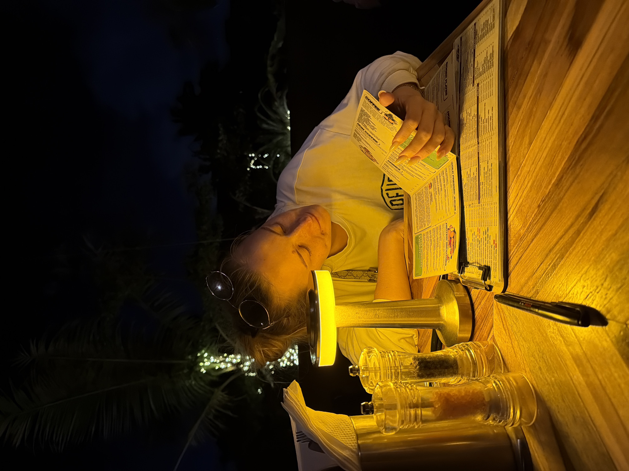 A woman is seated at a wooden table in what appears to be an outdoor restaurant setting at night. She is wearing a white sweatshirt and has round sunglasses pushed up onto her head. Her eyes are closed, and she is leaning forward, appearing to be tired or asleep, with her face illuminated by a small, yellow table lamp. In front of her, on the table, are menus, a pen, two clear salt and pepper grinders, and a metal container with napkins and cutlery. The background is dark, with some illuminated string lights visible among palm trees, suggesting a tropical environment, likely in the city of Mengwi, Indonesia.
