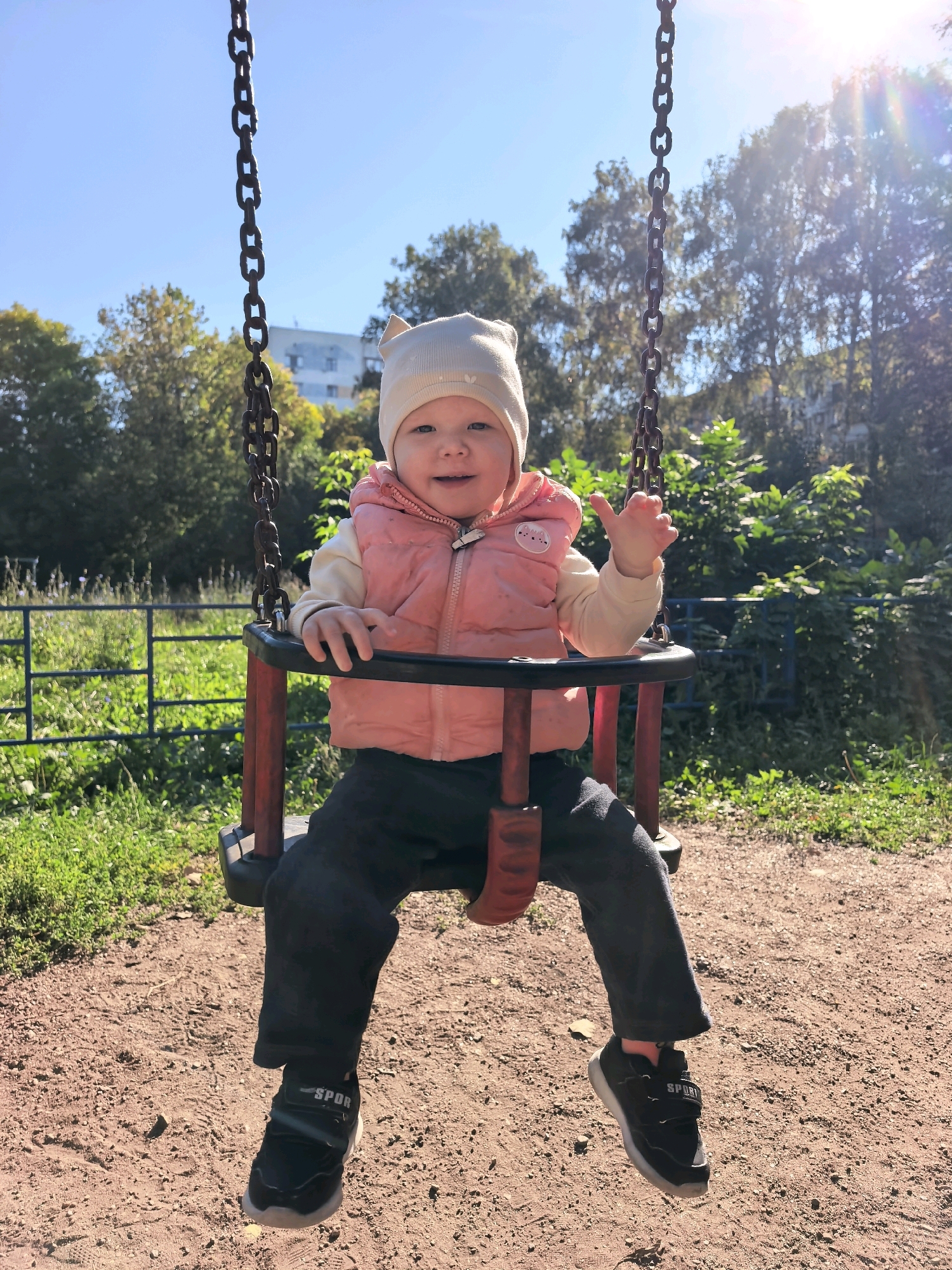 A baby wearing a light-colored hat with cat ears and a pink puffer vest is sitting on a swing in a playground. The baby is smiling and waving with their right hand. The swing has red metal supports and a black seat. The ground is covered with gravel. In the background, there are trees and a fence. The sky is clear and blue, with the sun shining brightly, causing lens flare. The location is identified as a playground in Sterlitamak, Russia.